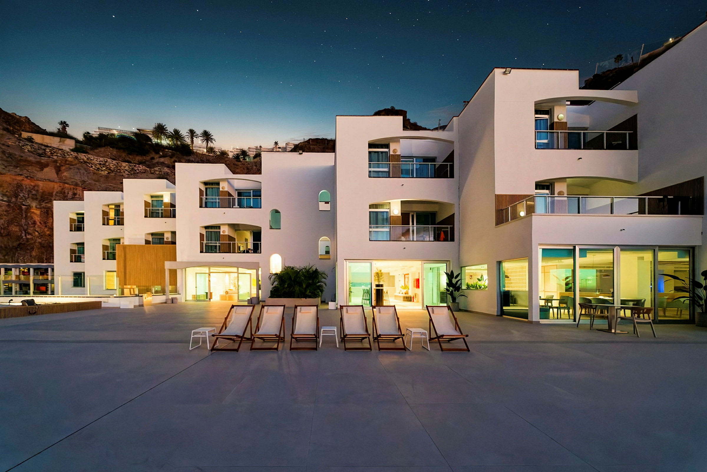 Bilde av en moderne hvit bygning med balkonger, stoler og bord på en terrasse mot en klippe ved solnedgang. (AI-generert tekst)