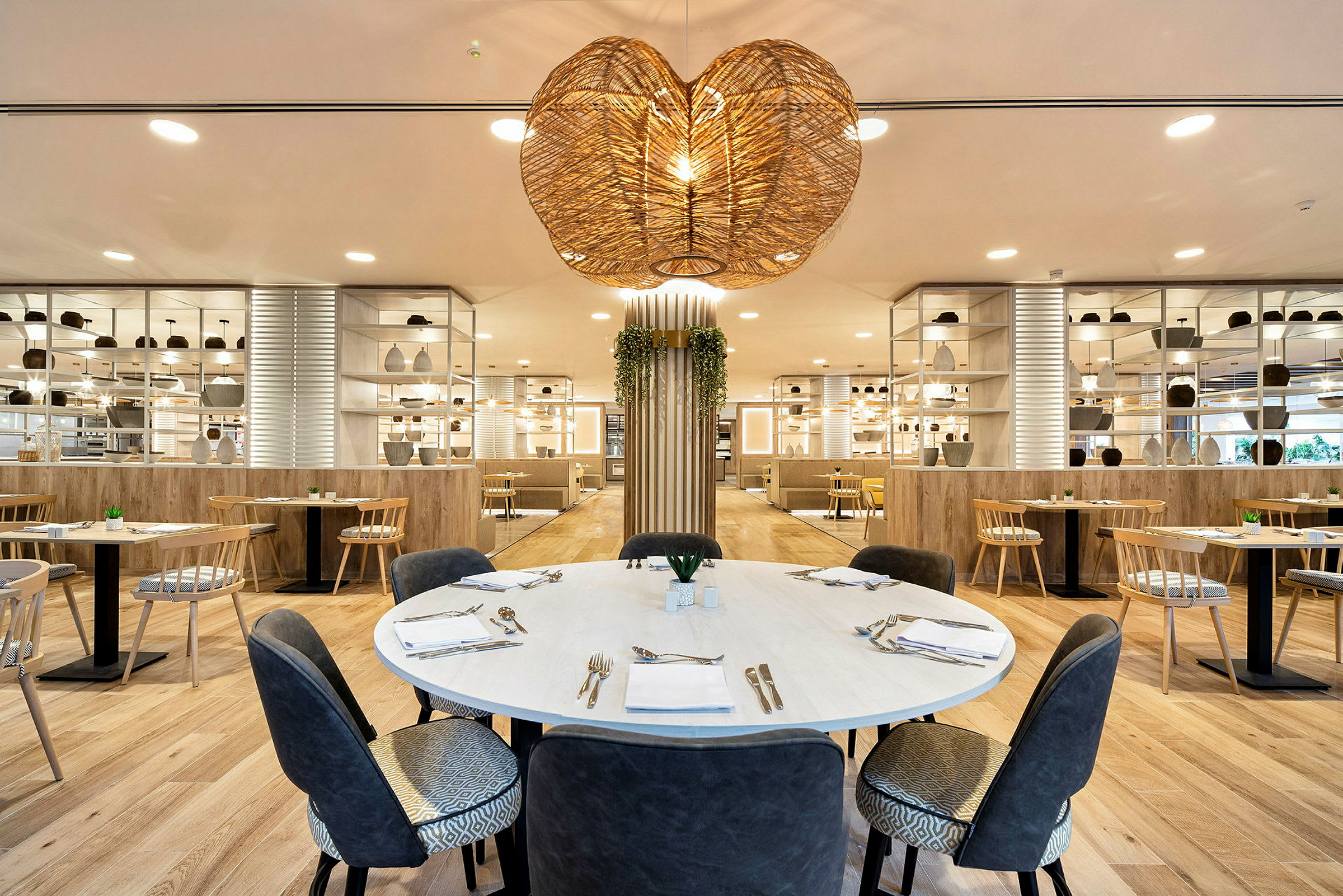 Et moderne innredet restaurantinteriør med et rundt bord i forgrunnen, omgitt av sitteplasser og dekorative hyller. (AI-generert tekst)