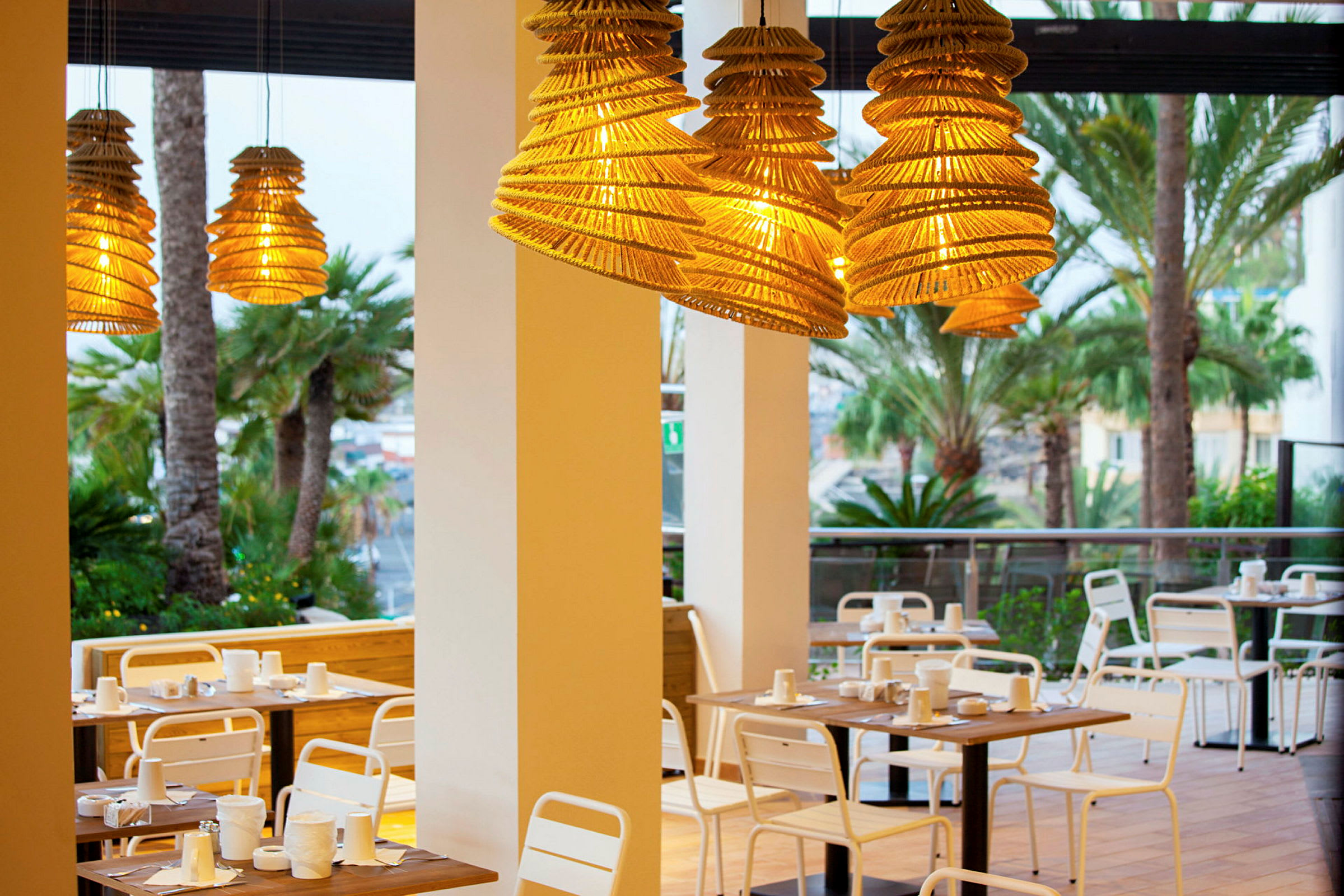 Utemøbler ved restaurant med hengende lampeskjerm av tre og palmetrær i bakgrunnen. (AI-generert tekst)