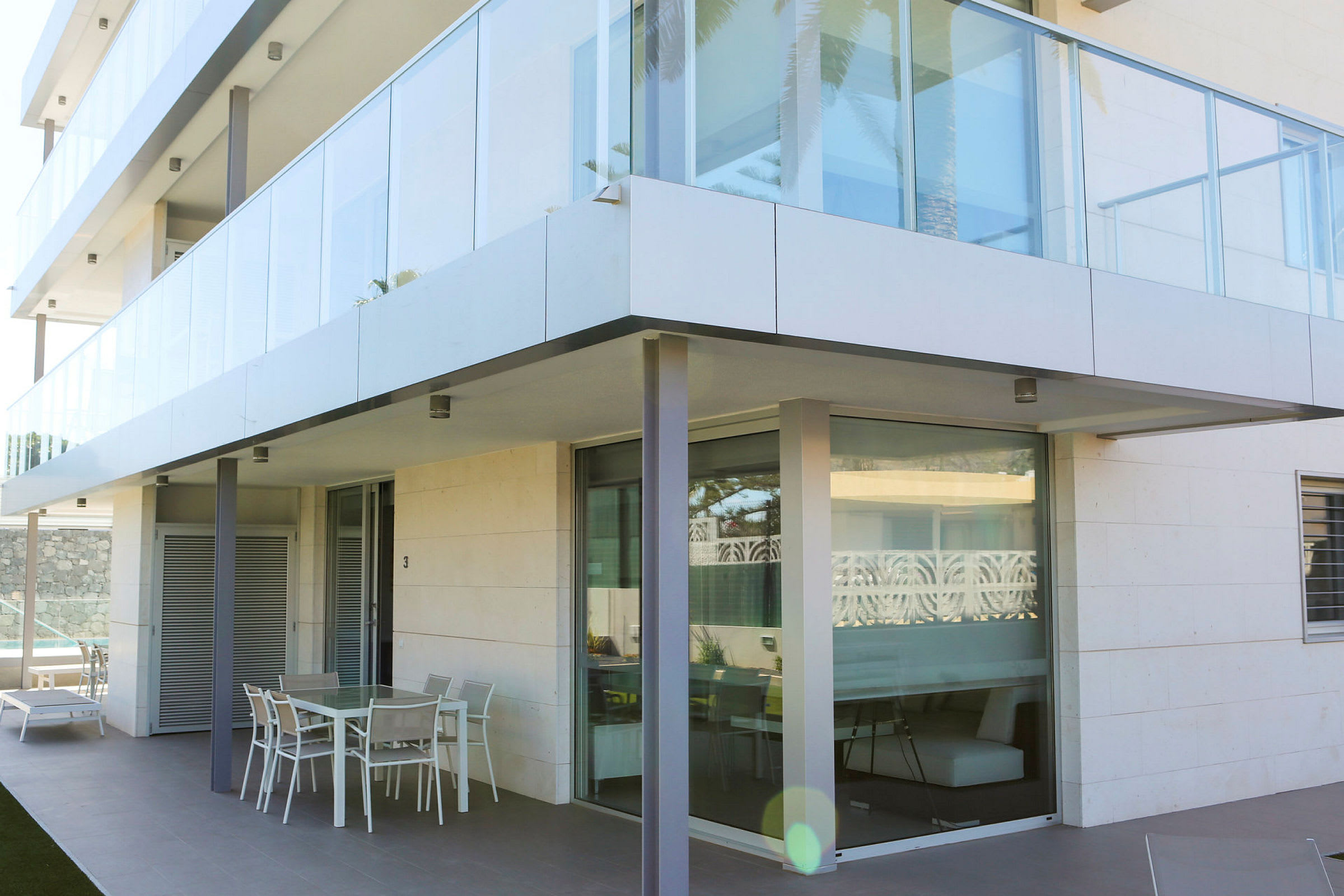 Det moderne flerfamiliehuset har en glassfasade med en utendørsterasse i første etasje som inkluderer utemøbler. (AI-generert tekst)