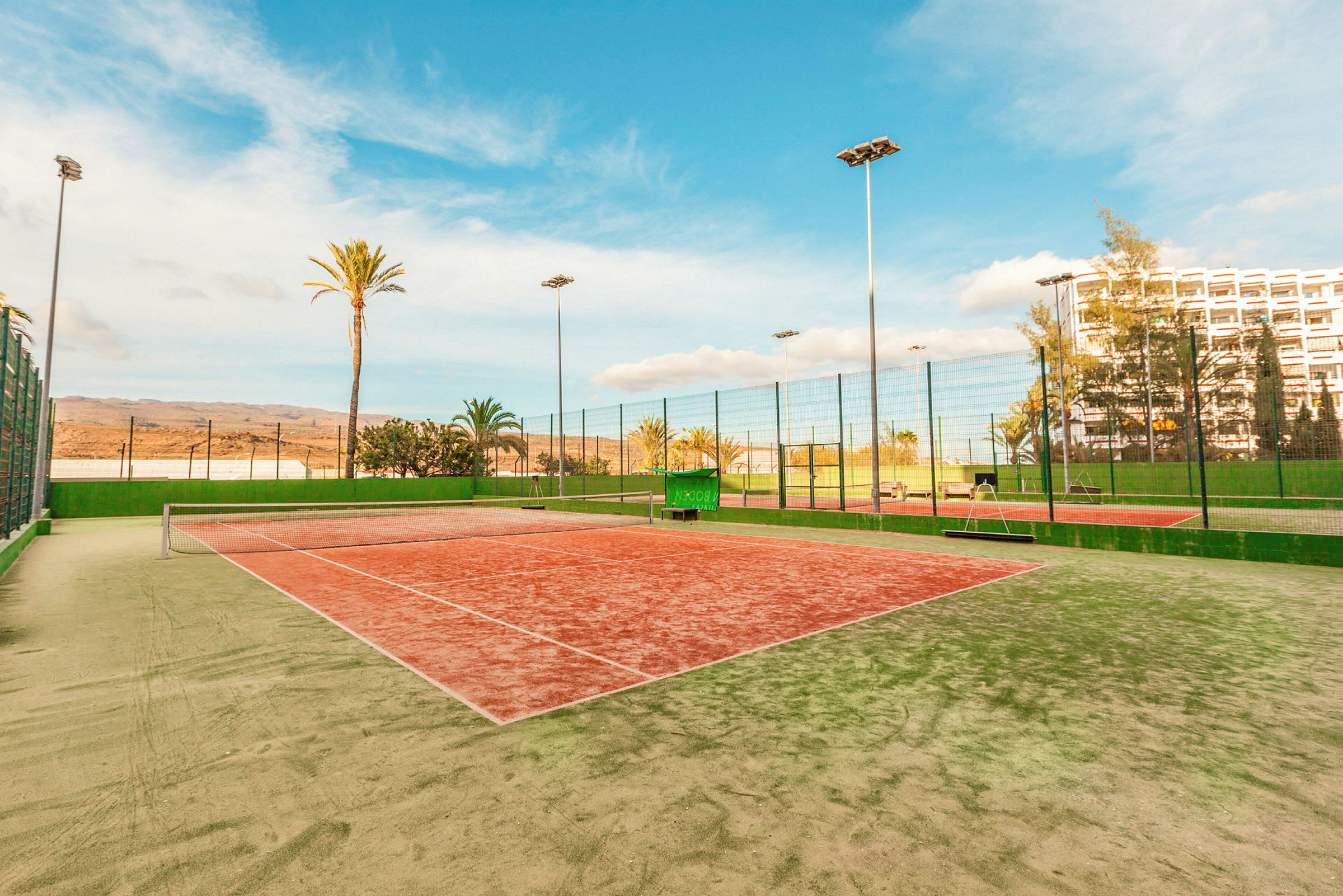 En tennisbane med grønn og rød overflate, omgitt av høye nett og palmer. (AI-generert tekst)