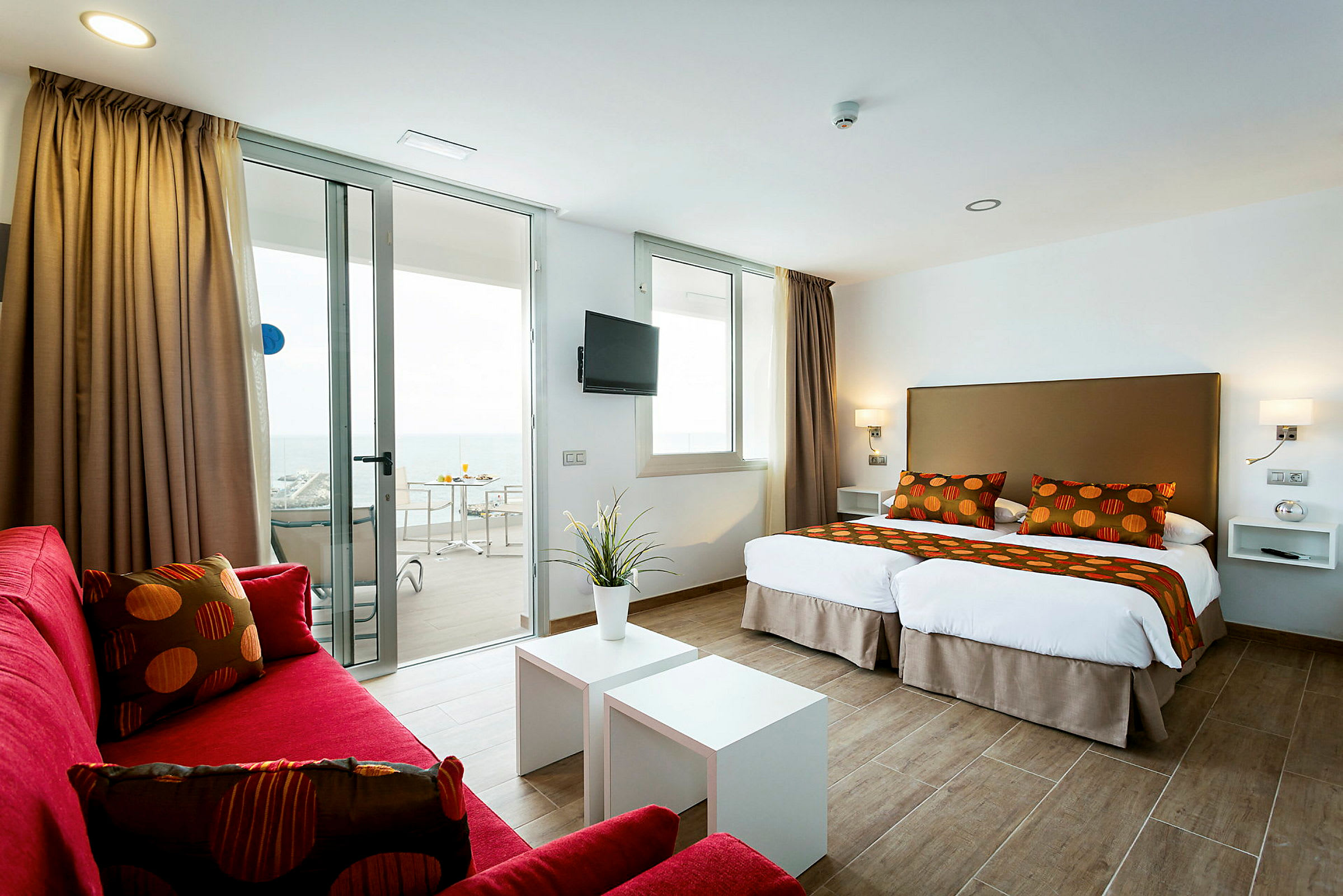 Et moderne hotellrom med to enkeltsenger, røde sofaer og en balkong med havutsikt. (AI-generert tekst)