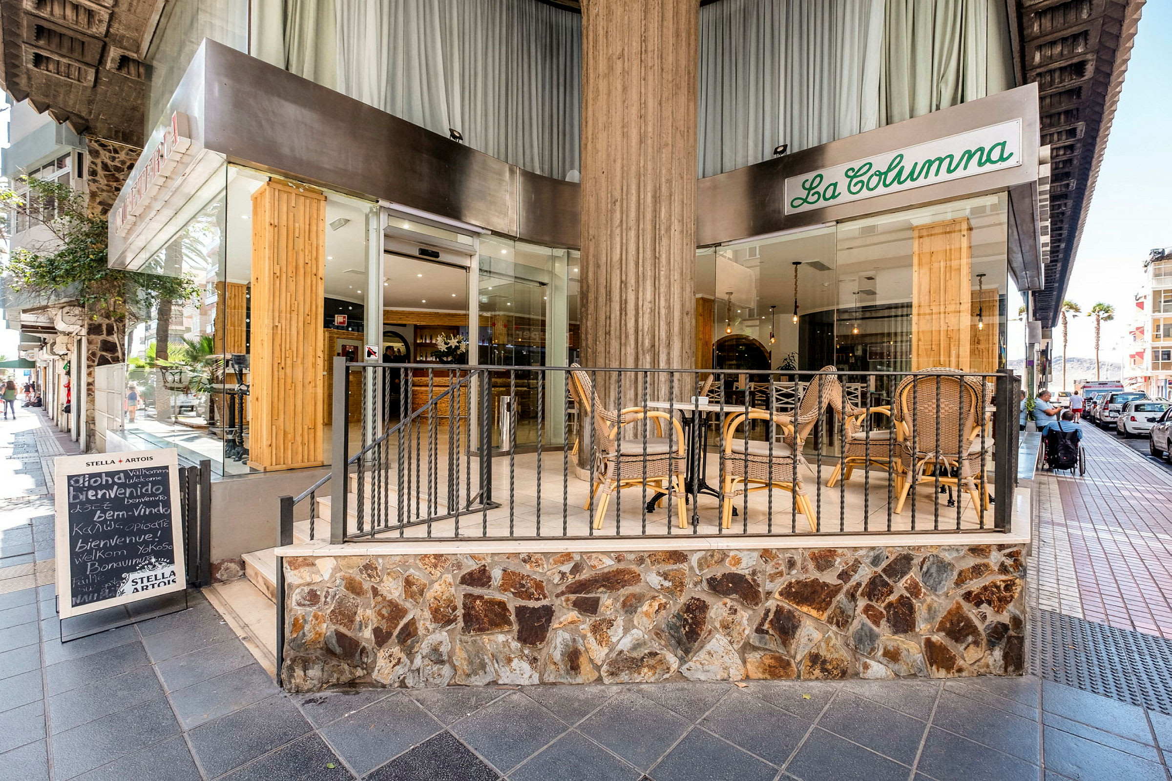 Inngangen til en restaurant med uteservering hvor det står bord og stoler. Restauranten heter "La Columna" og ligger ved et fortau. (AI-generert tekst)