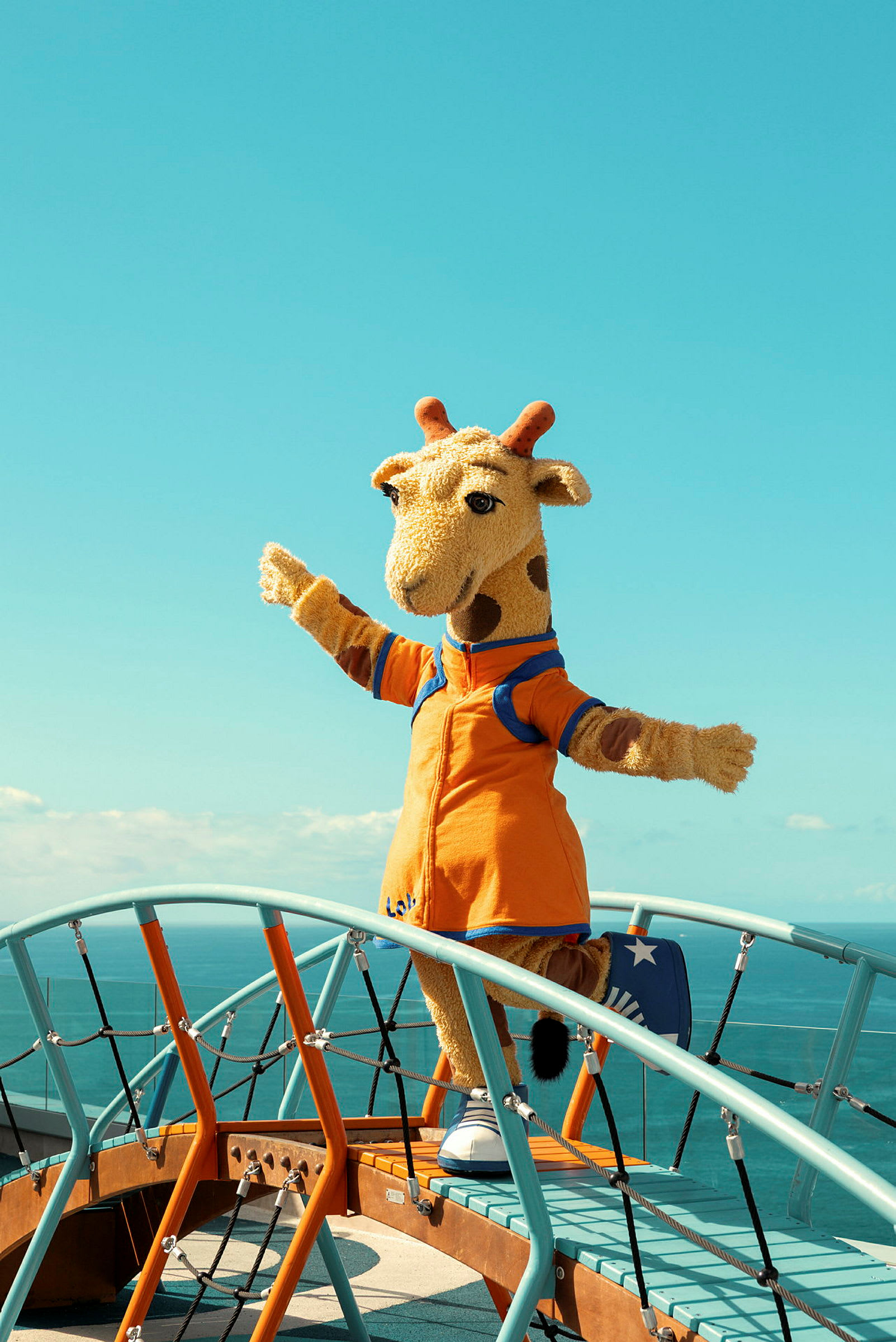 En person kledd som en giraffmaskot står på en lekeplass med havet i bakgrunnen. (AI-generert tekst)