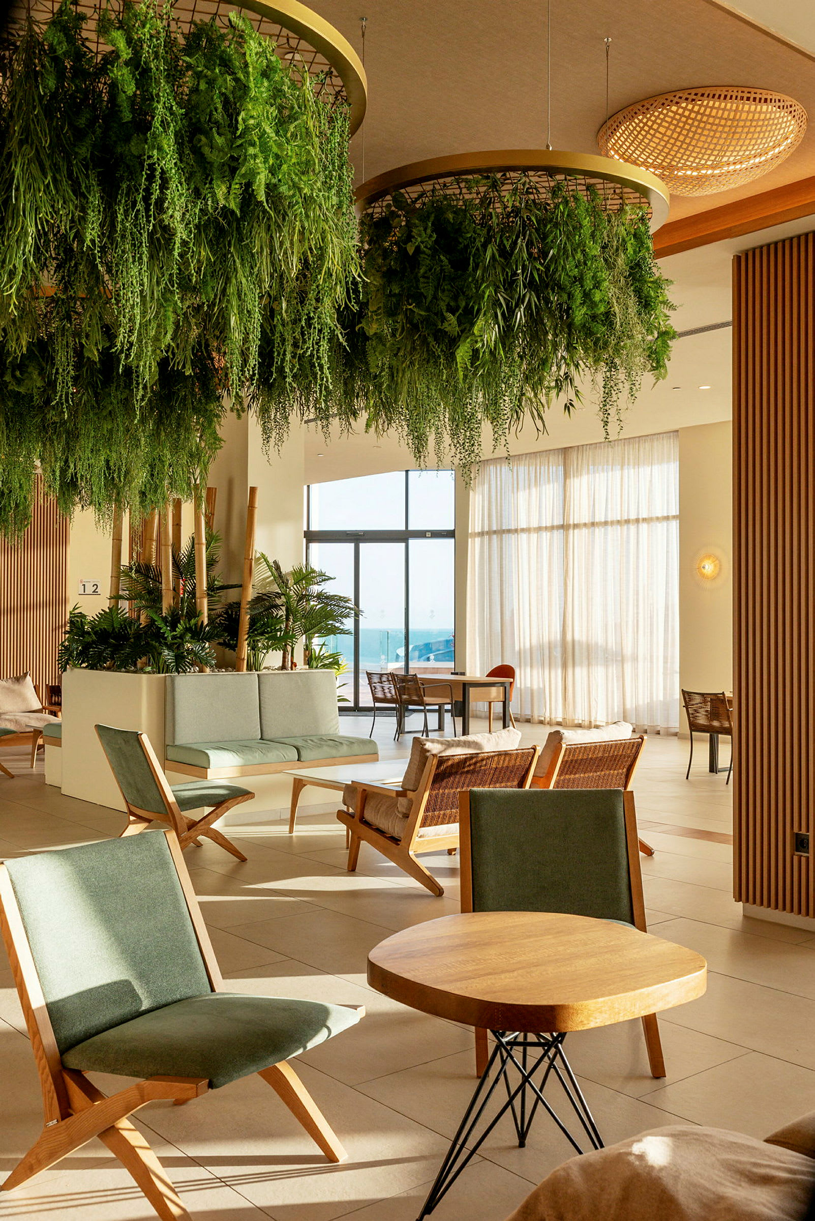 En hotellobby med grønne planter hengende fra taket, sittegrupper med trestoler og bord, og glassdører som fører ut til en solrik terrasse. (AI-generert tekst)