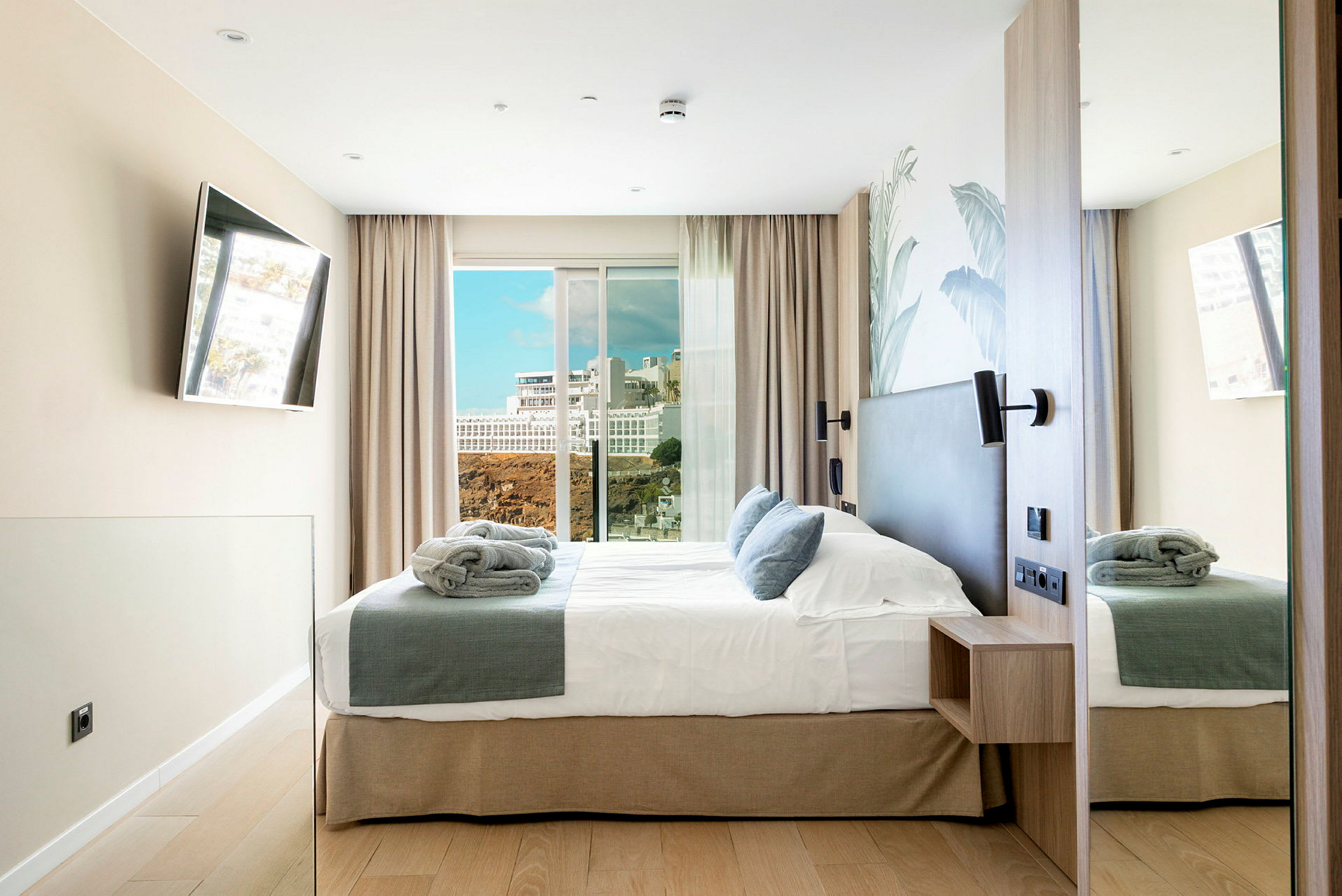 Et hotellrom med en dobbeltseng, lyst innredede vegger, vindu med utsikt over en bygning og klipper. (AI-generert tekst)