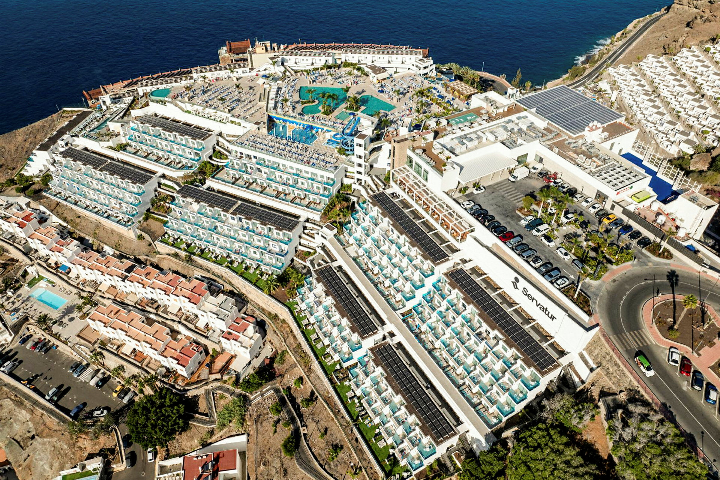 Luftfoto av et stort hotellkompleks ved havet med bassengområde og omkringliggende bolighus. (AI-generert tekst)