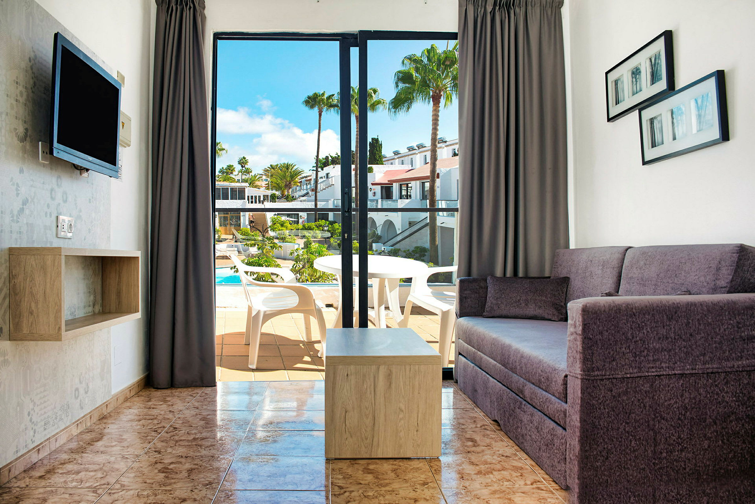 Utsikt over en stue med sofa og bord samt en åpning til en balkong med utsikt over et basseng. (AI-generert tekst)