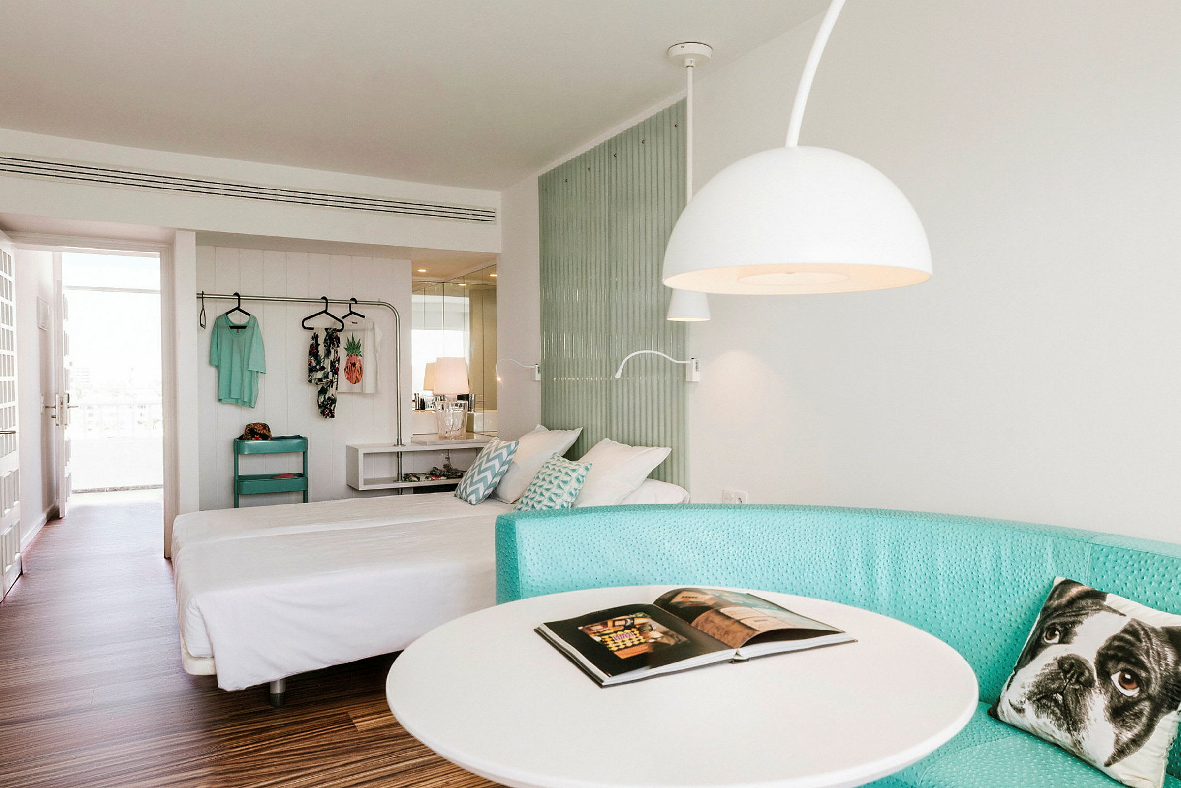 Et hotellrom med en dobbeltseng, turkis sofa, et rundt bord og en hengende klesholder. (AI-generert tekst)
