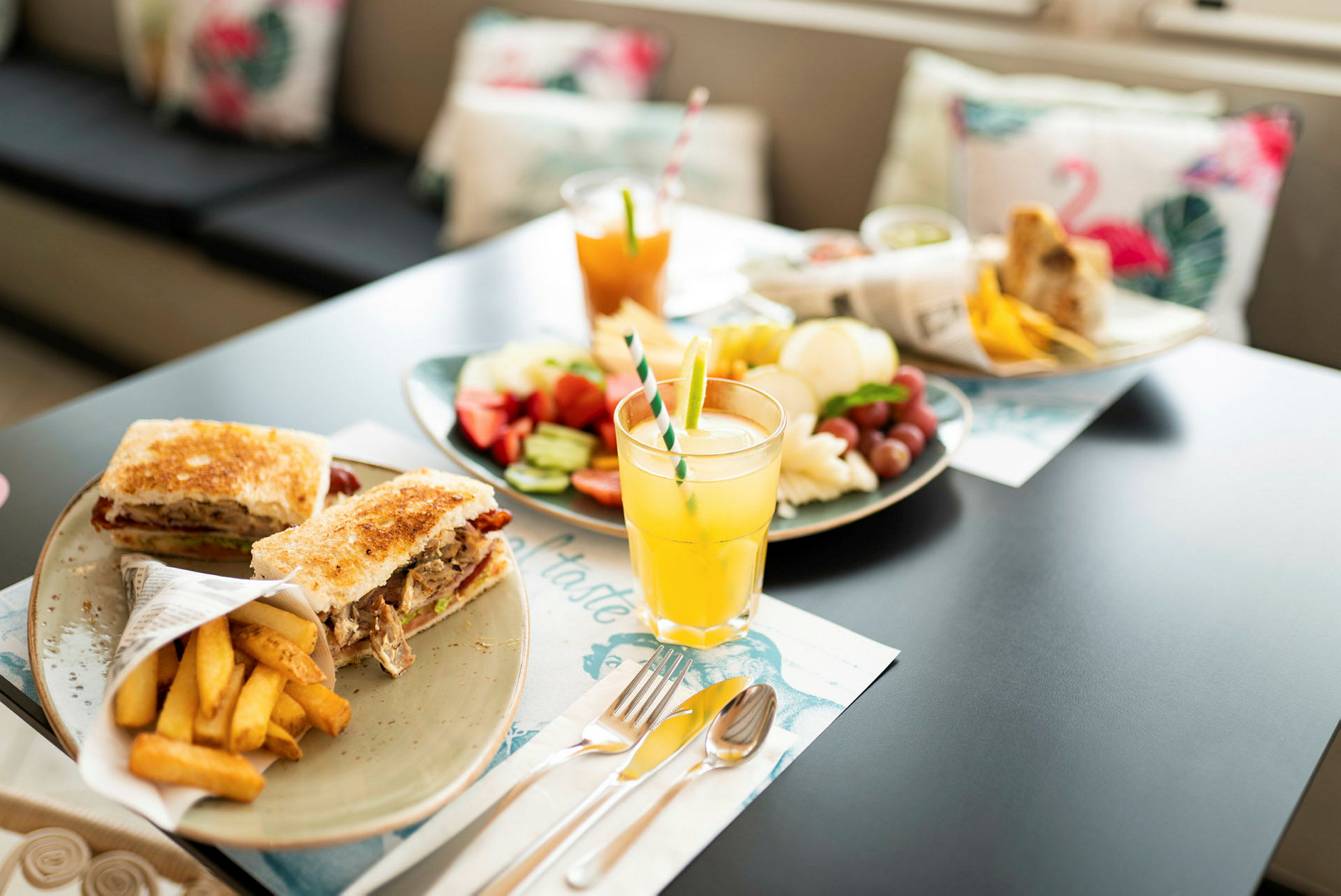 Et bilde av et bord med forskjellige tallerkener som inneholder mat og drikke, inkludert smørbrød, pommes frites, frukt og juice. (AI-generert tekst)