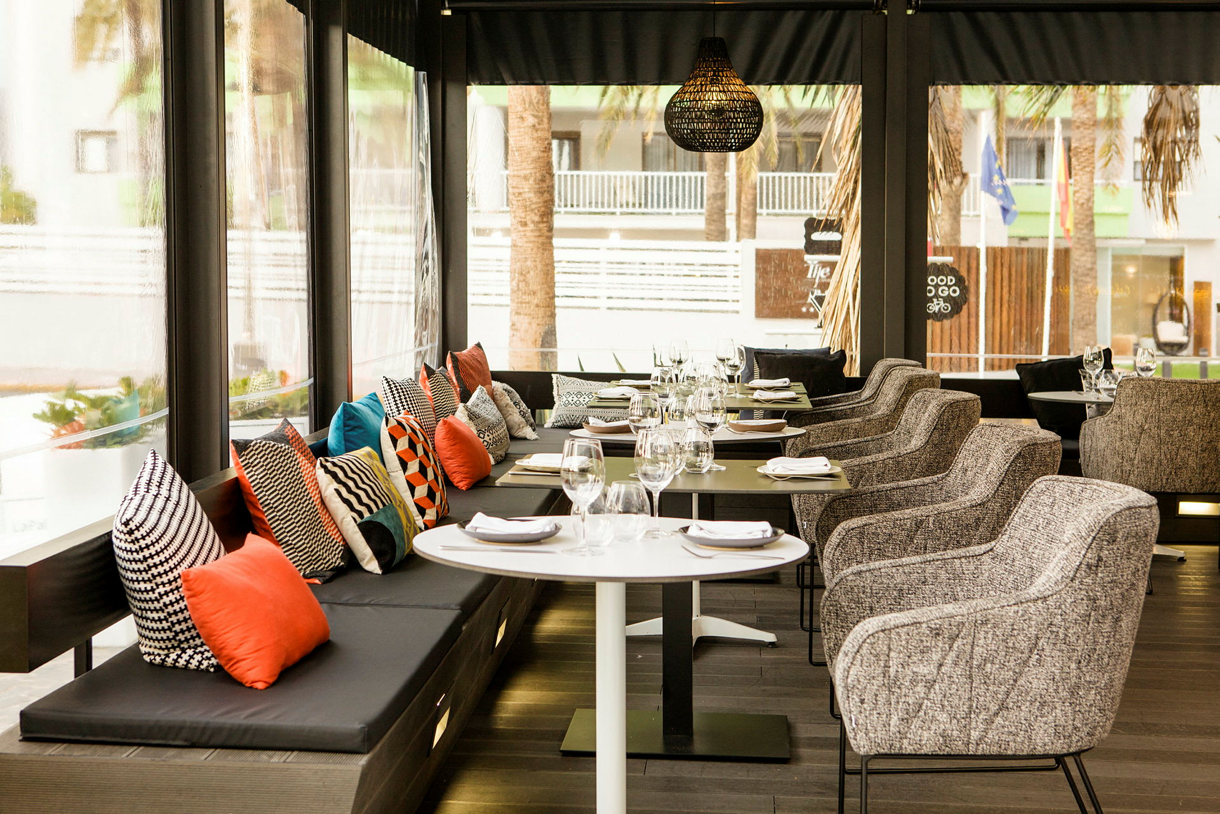 En restaurant med bord og stoler, samt en benk med fargerike puter. (AI-generert tekst)