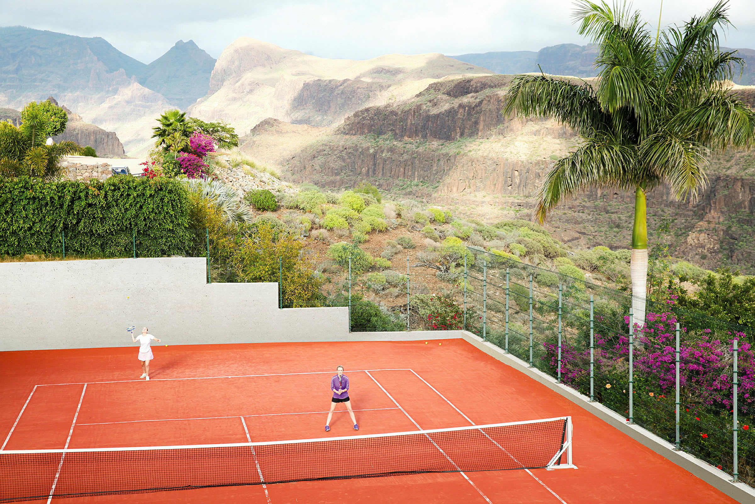 To personer spiller tennis på en rød tennisbane, omgitt av en grønn dal. (AI-generert tekst)
