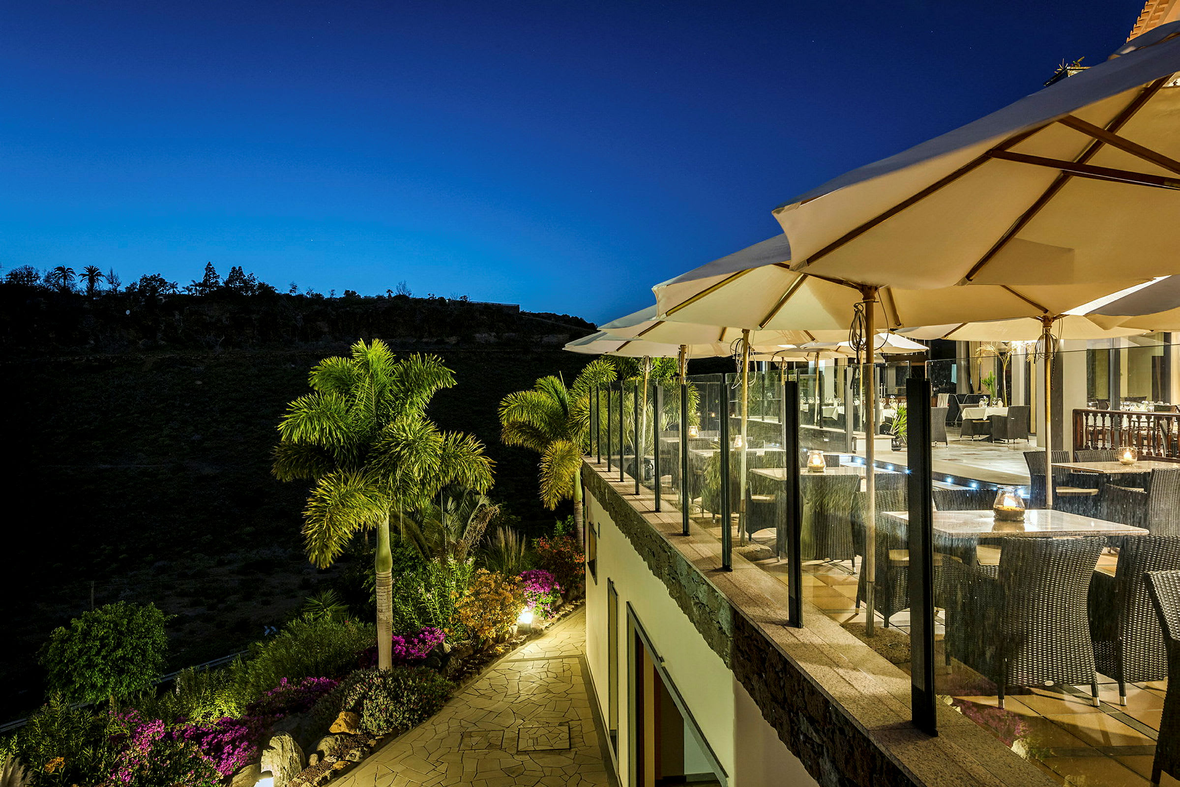 Restaurantterrasse med parasoller og bord, omgitt av grøntområder, om kvelden. (AI-generert tekst)