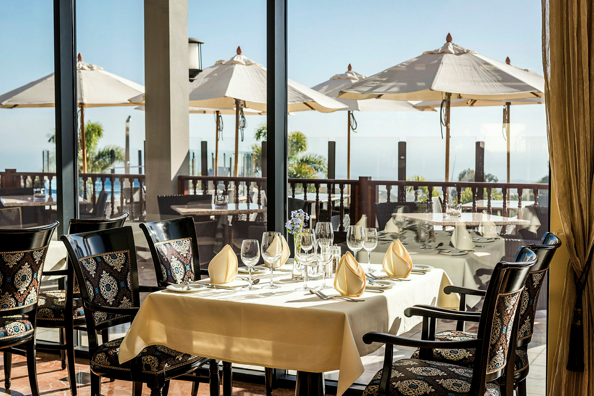 Et dekket bord i en restaurant med utsikt over en terrasse med parasoller og havet. (AI-generert tekst)