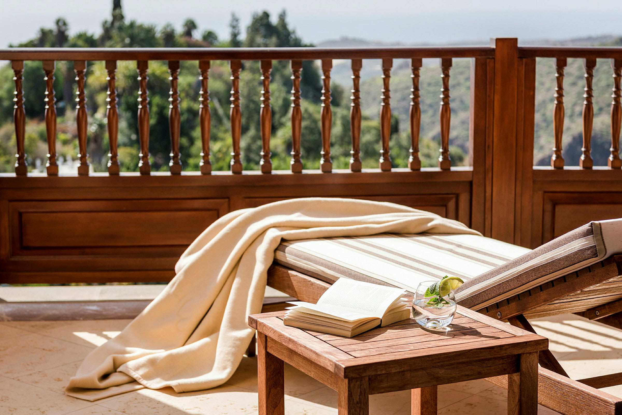 Solstol og trebord med bok og drink, terrasse med trebalustrade og utsikt. (AI-generert tekst)