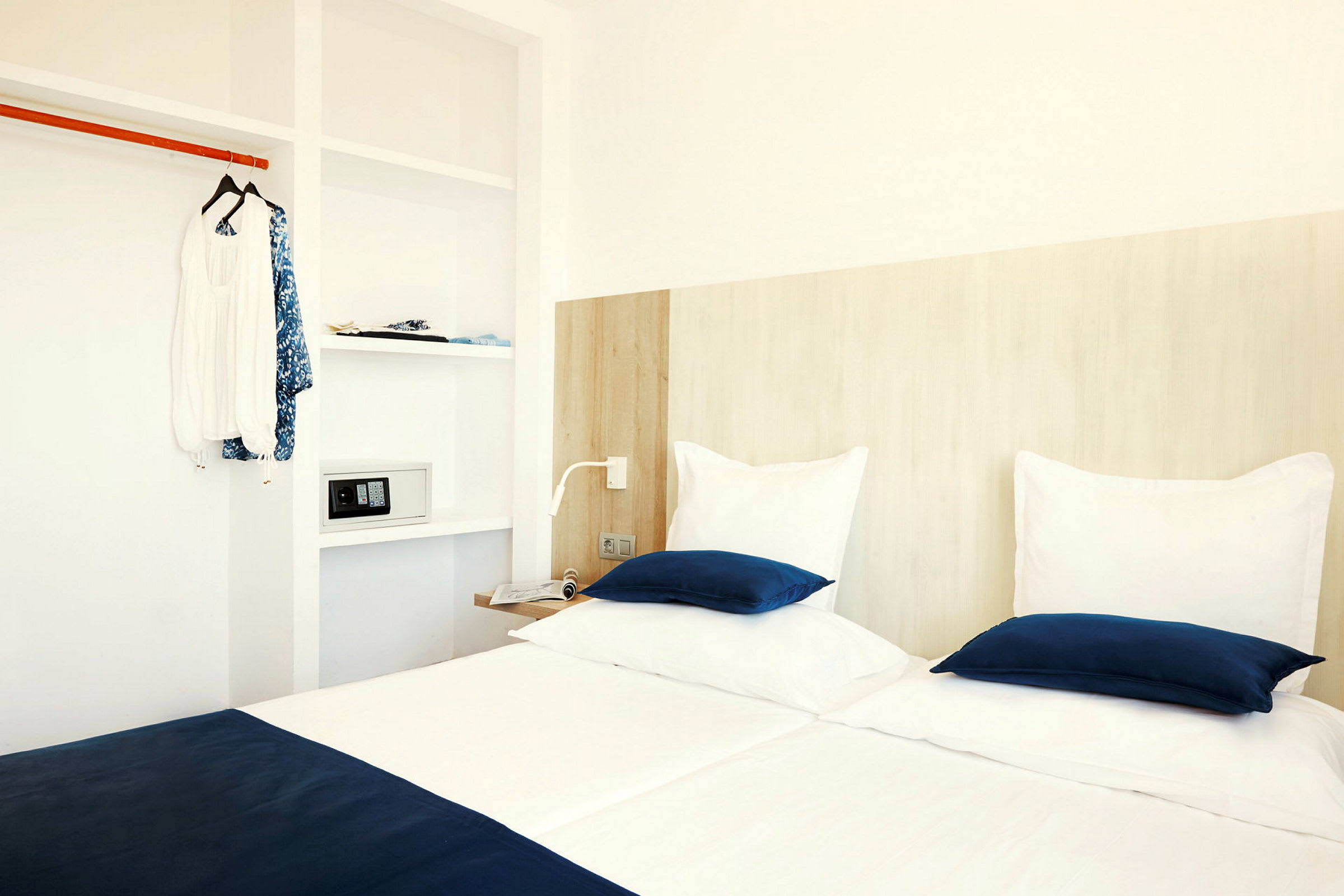 Et hotellrom med to senger med hvite og blå sengetøy, en garderobe med klær og en safe. (AI-generert tekst)