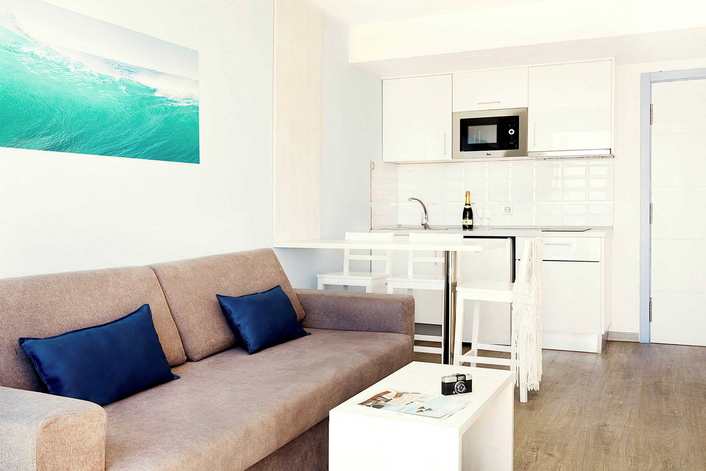 En lys og moderne stue med en sofa, to puter, et lite bord, samt et kjøkken med hvit innredning i bakgrunnen. (AI-generert tekst)
