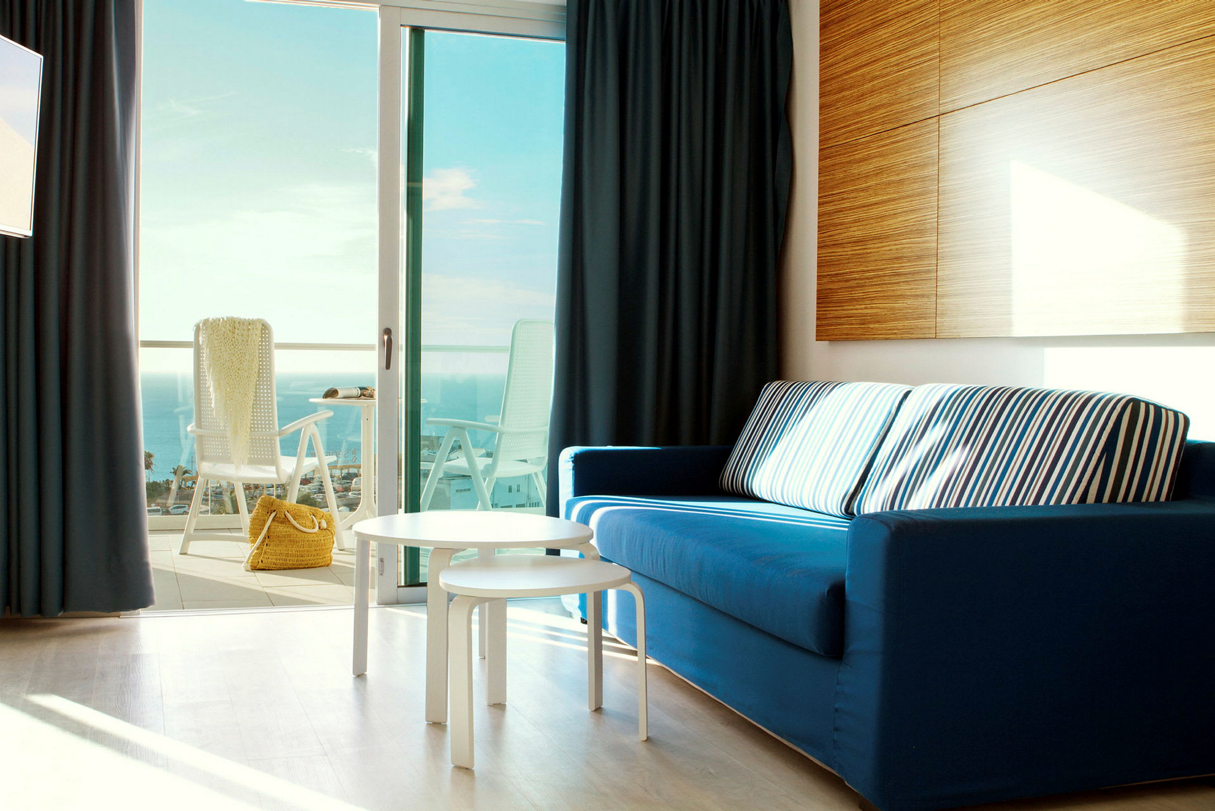 Et interiørbilde med en blå sofa, hvite bord og en åpen balkongdør med utsikt over havet. (AI-generert tekst)