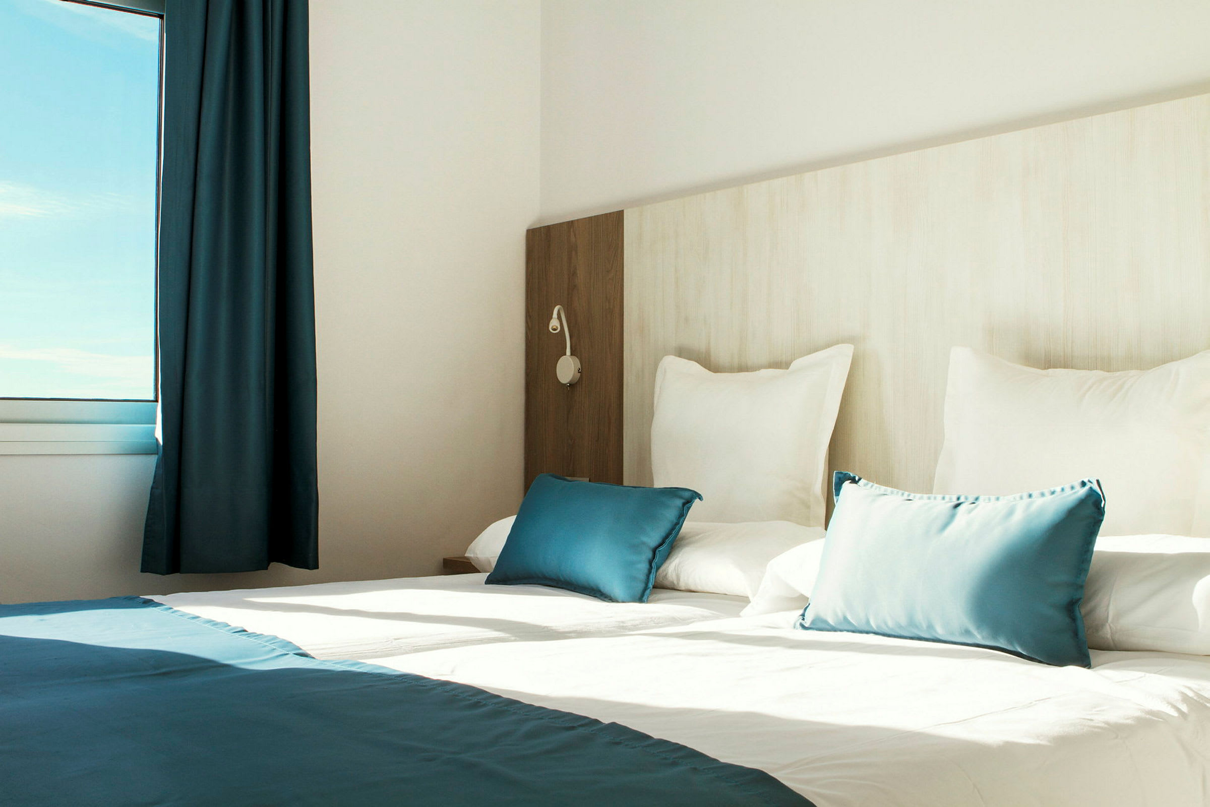 Interiør av et lyst hotellrom med en oppredd seng, hvite og blå puter samt et vindu som slipper inn naturlig lys. (AI-generert tekst)