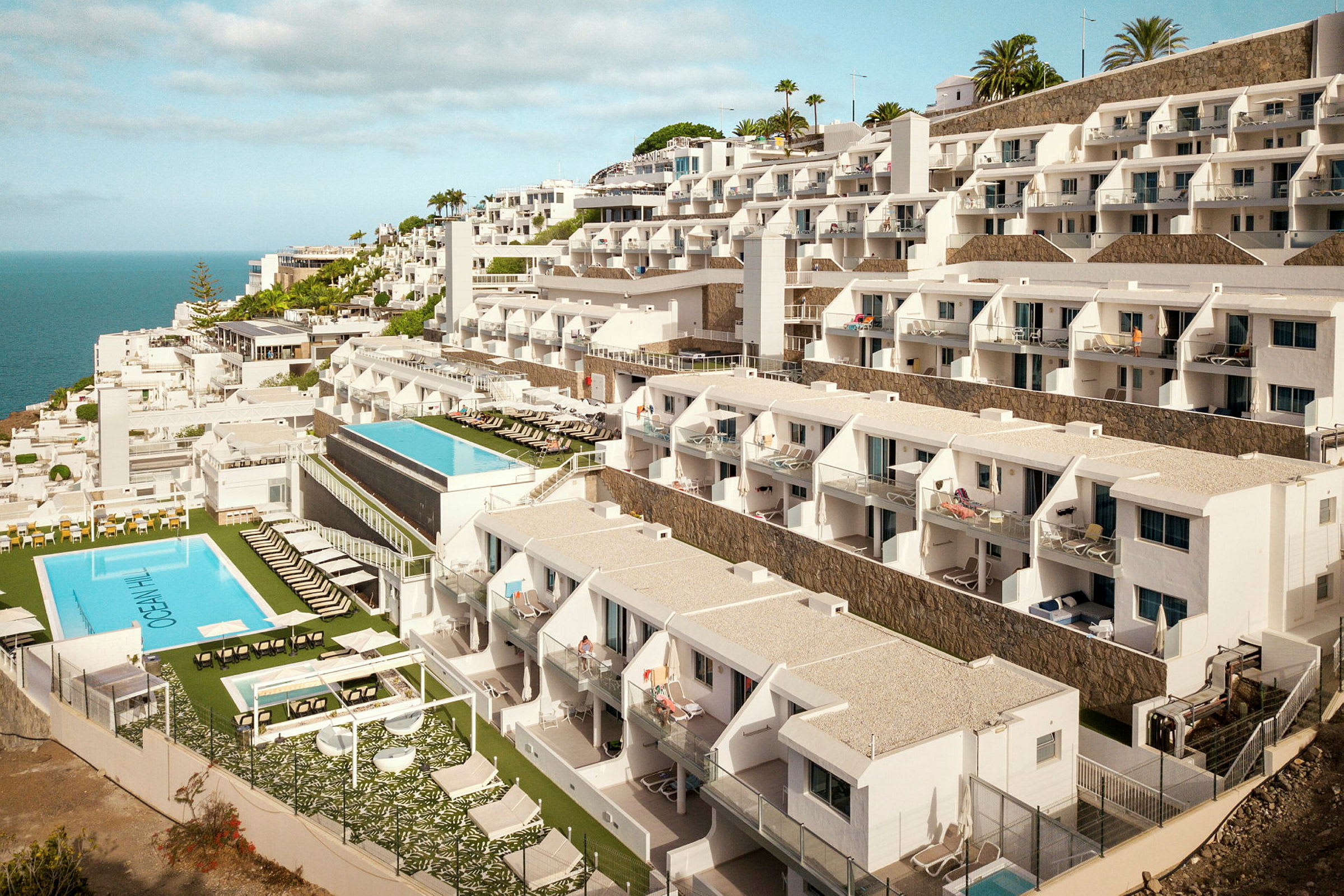 Hotellbygning med flere etasjer og bassenger mot havet. (AI-generert tekst)