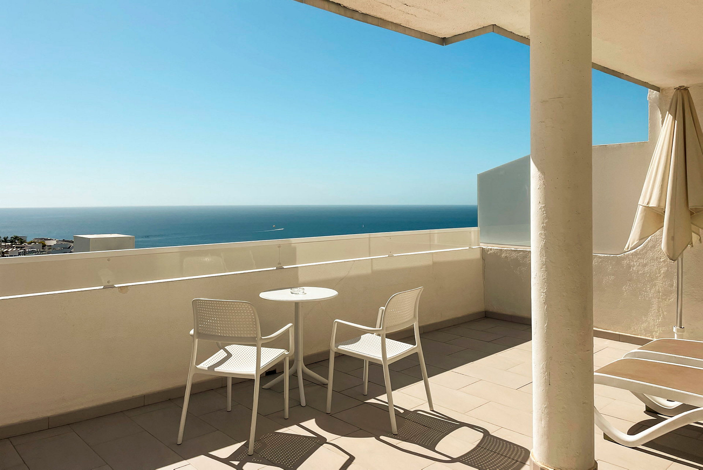 Terrasse med to hvite stoler og et bord, havet er synlig i bakgrunnen. (AI-generert tekst)