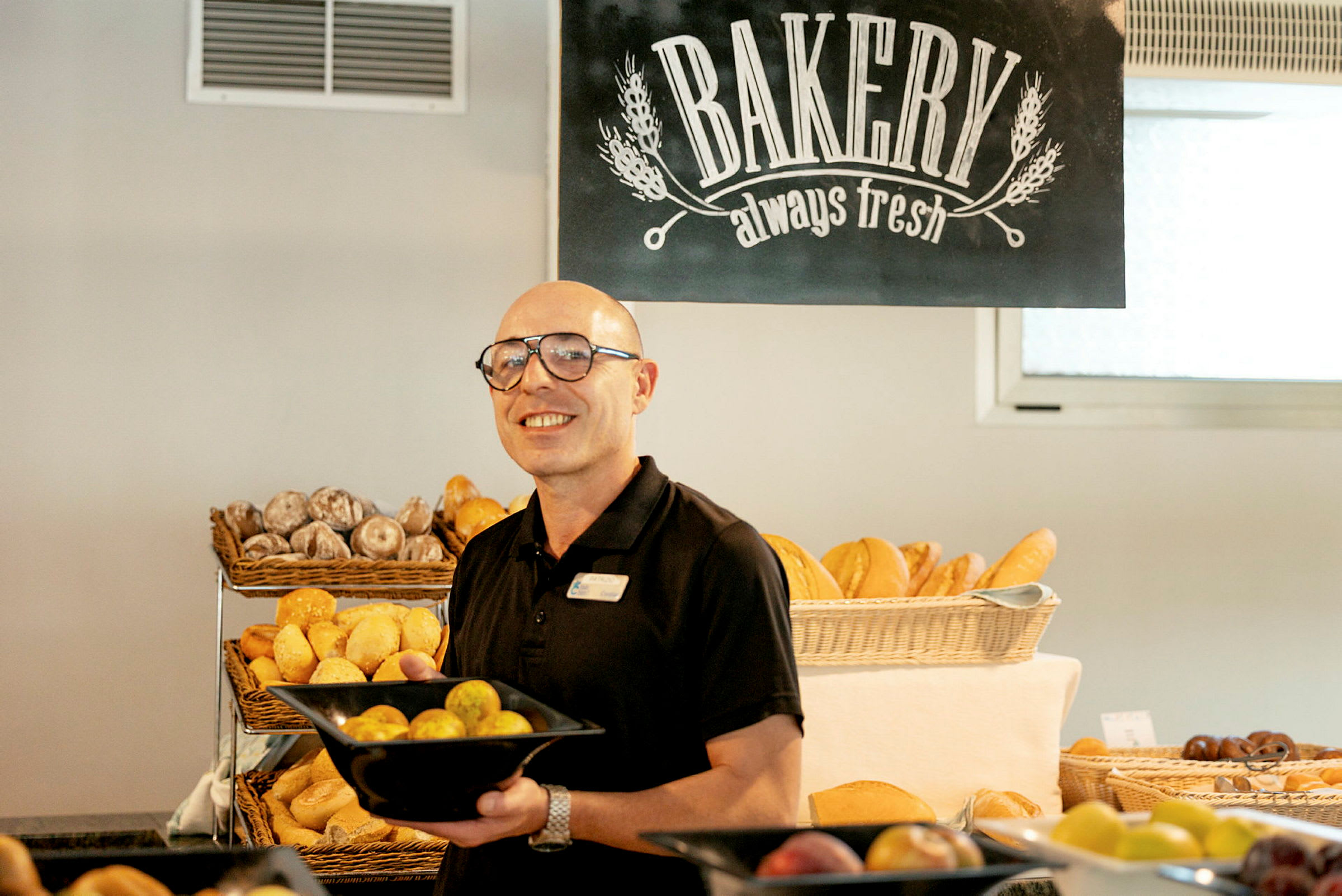 En person holder en svart bolle fylt med brød i et bakeri, med andre bakverk og et skilt med teksten 'Bakery always fresh' synlige i bakgrunnen. (AI-generert tekst)