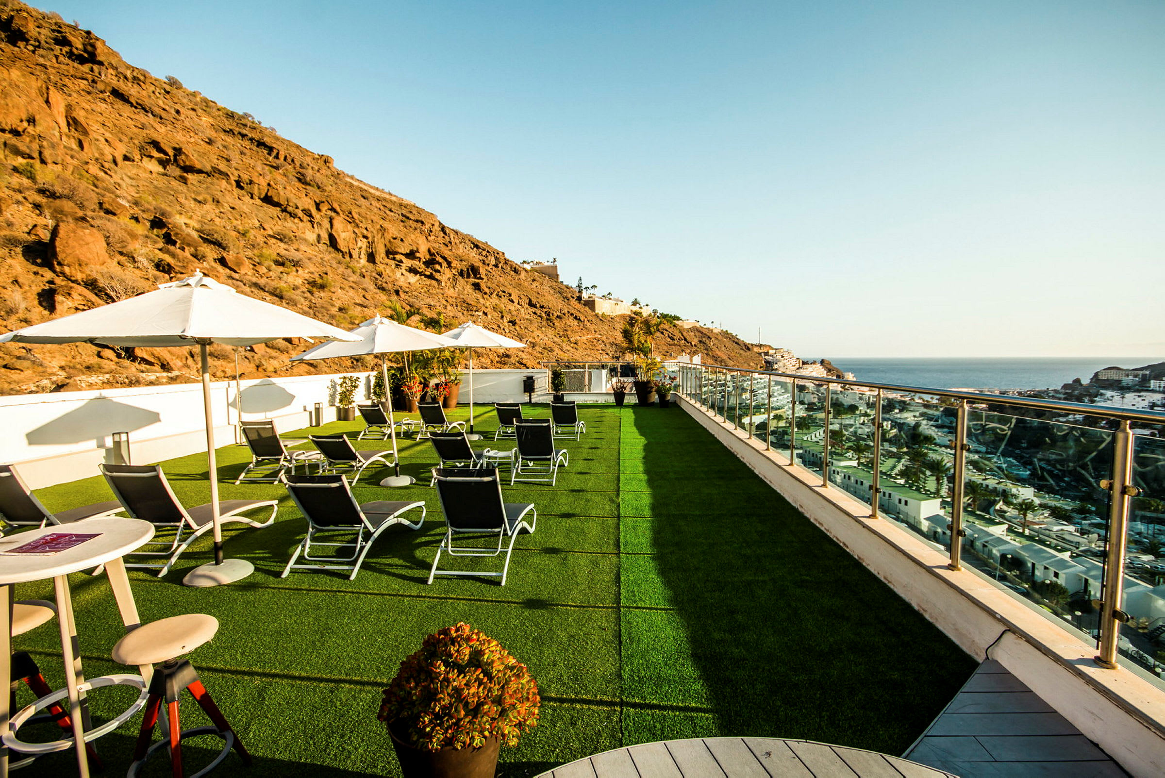 Bilde av en terrasse med solsenger, parasoller og utsikt over havet og omkringliggende klipper. (AI-generert tekst)