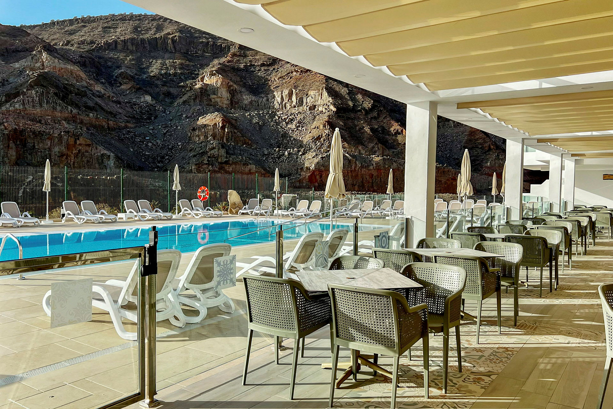 Utsikt over et basseng med solsenger og parasoller foran en fjellaktig bakgrunn samt spiseområde med bord og stoler på en terrasse. (AI-generert tekst)