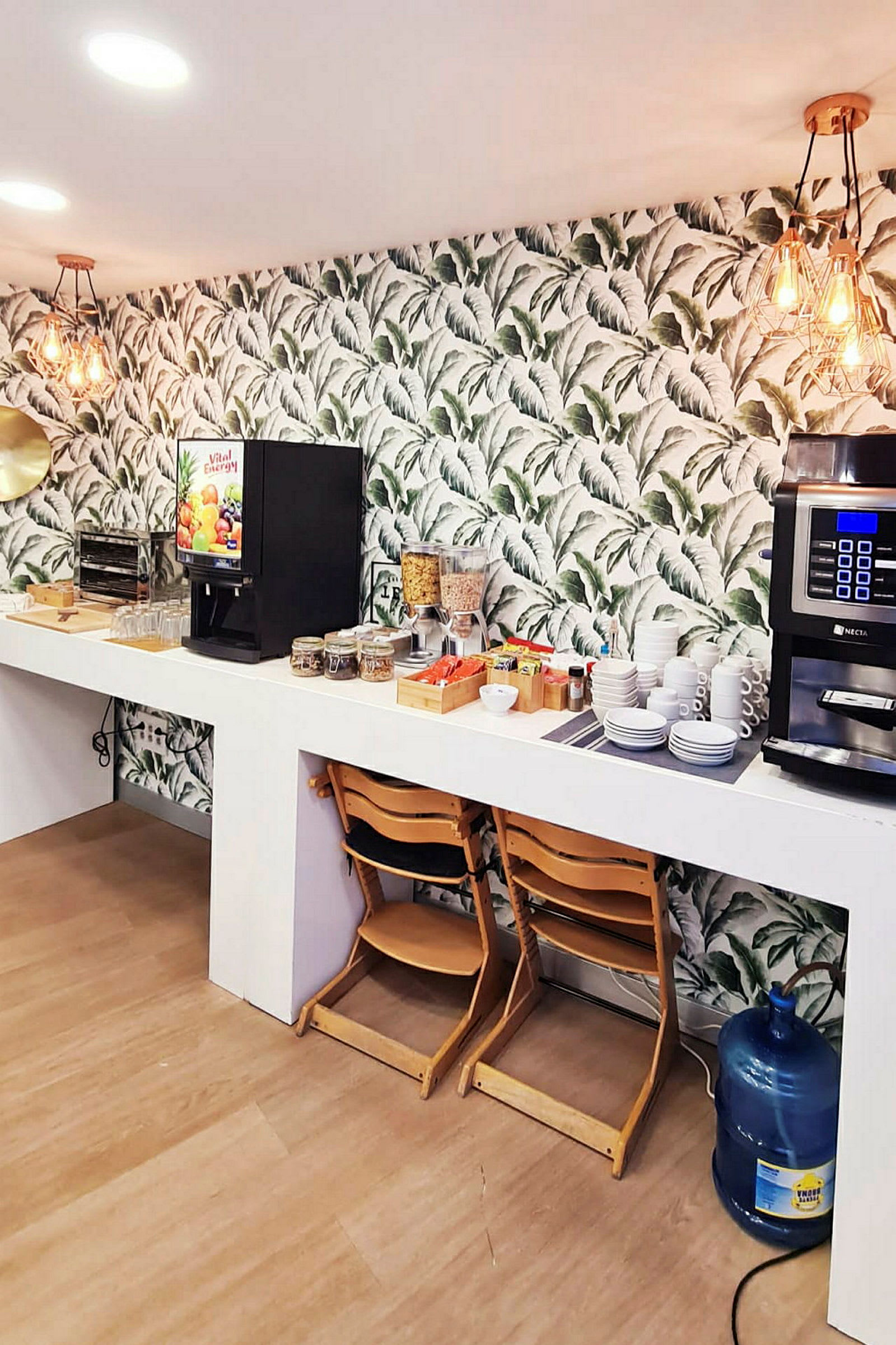 Bilde av en frokostbuffé med kaffemaskin, juiceautomat, tallerkener, kopper og matvarer presentert på en hvit kjøkkenbenk. Stol og vannbeholder i forgrunnen. (AI-generert tekst)