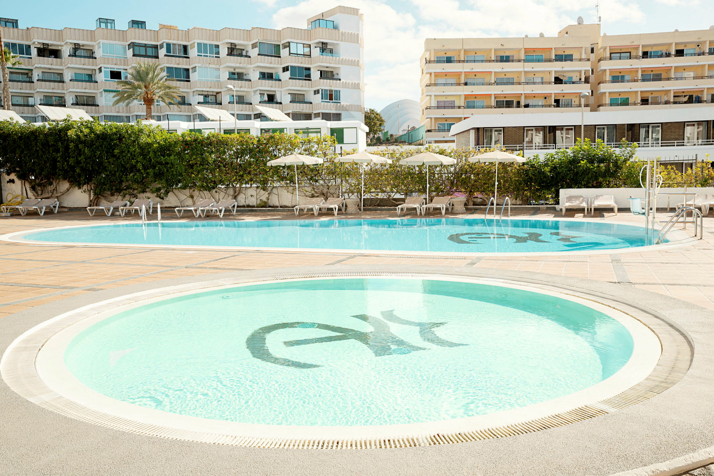 To svømmebassenger omgitt av solsenger og parasoller, med hotellbygninger i bakgrunnen. (AI-generert tekst)