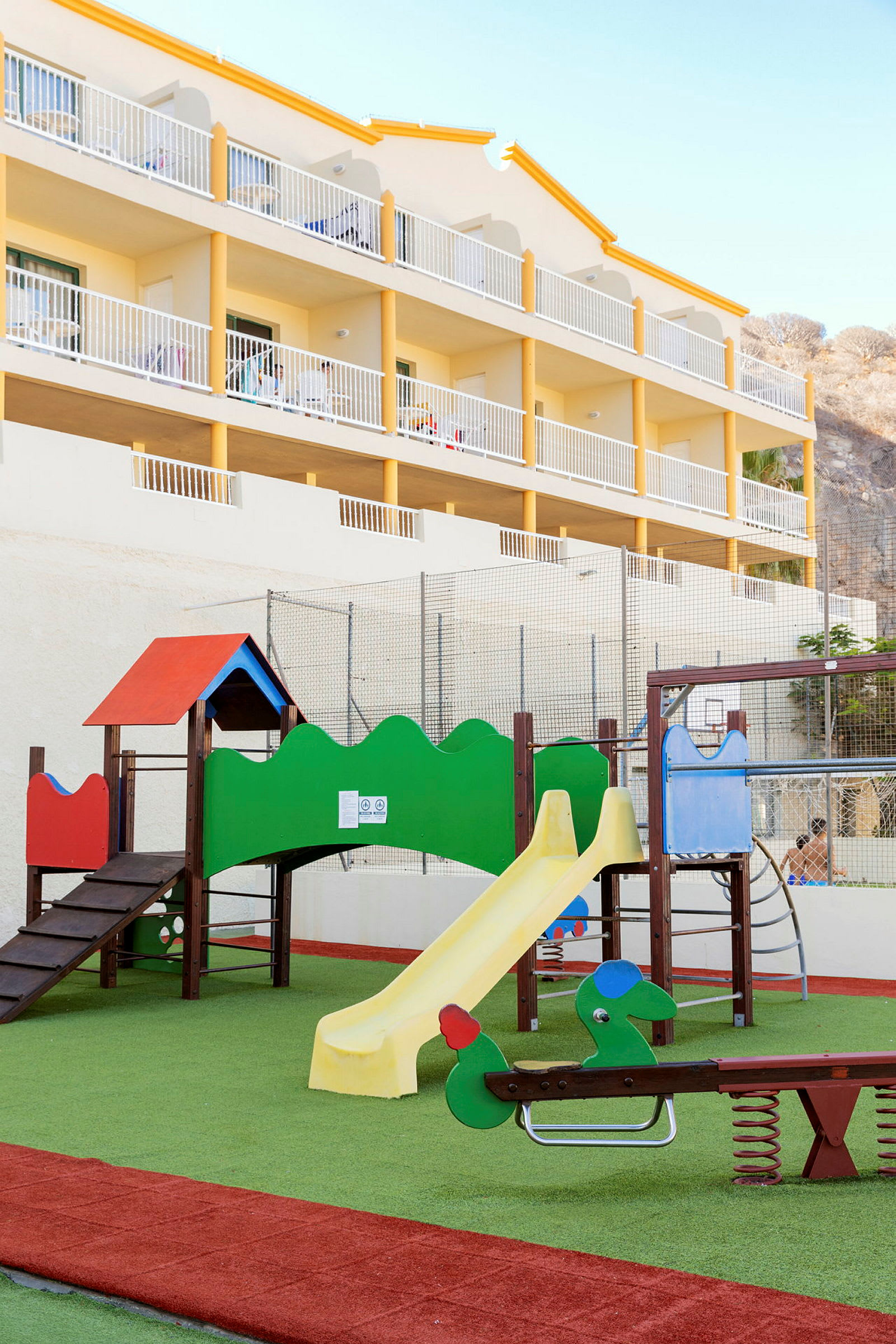Lekeplass med sklie og lekeutstyr foran en hotellbygning. (AI-generert tekst)