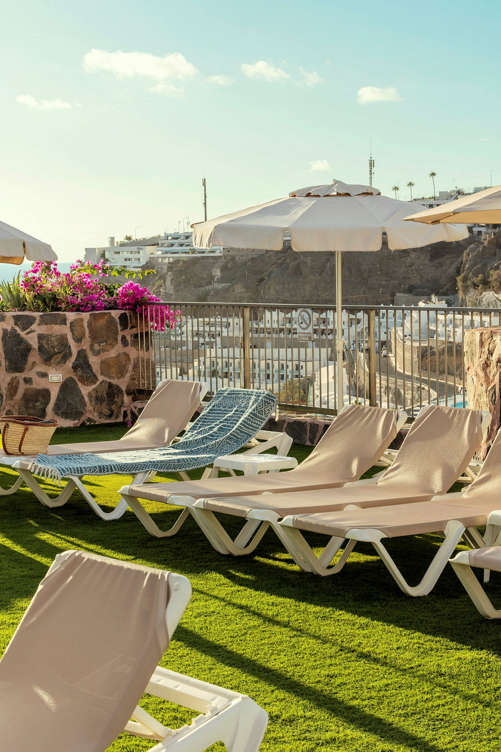 Bilde av en solterrasse med solsenger, parasoller og en blomsterpotte. I bakgrunnen sees et boligområde og åser. (AI-generert tekst)