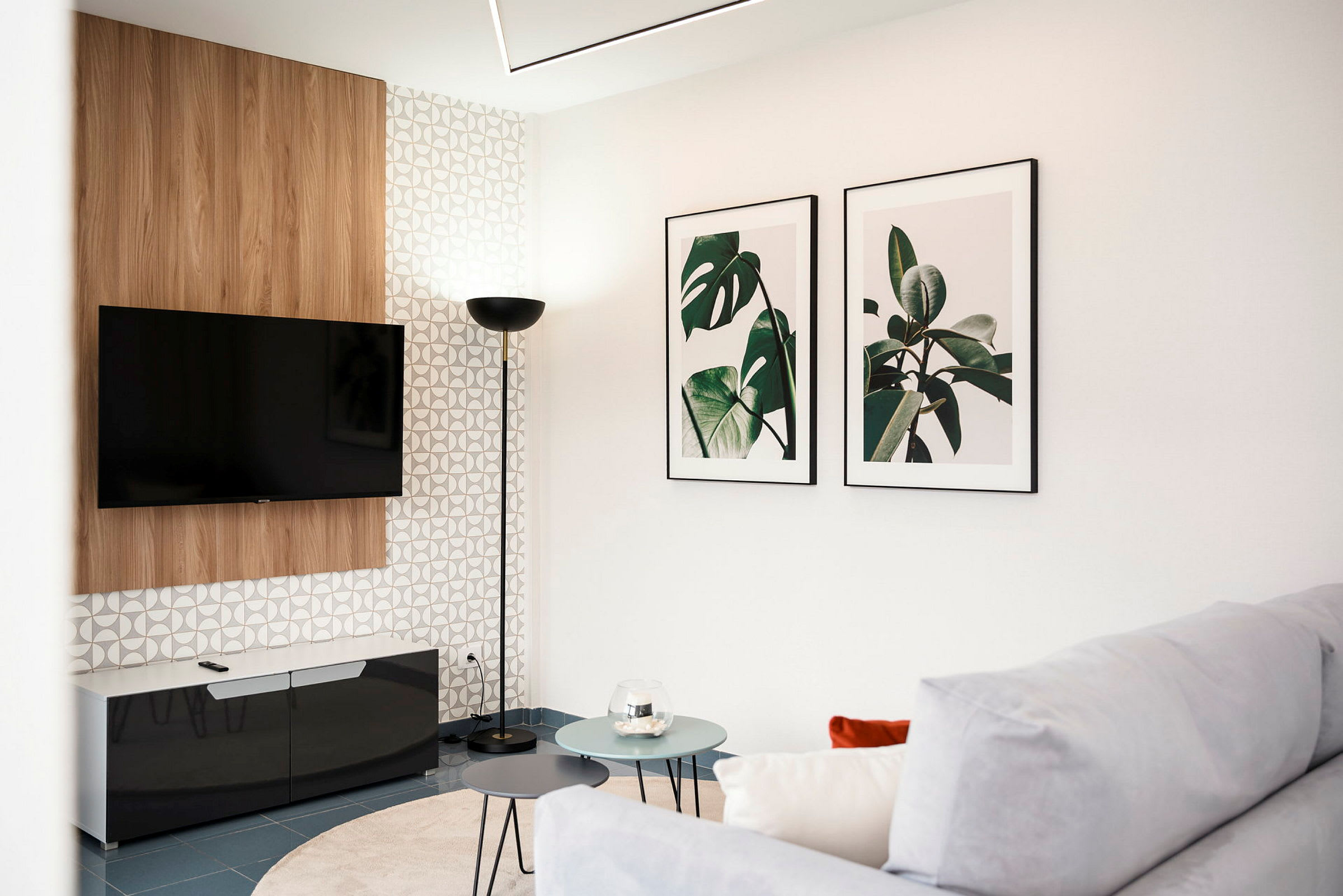 Et moderne stue med en veggmontert TV, to bilder med plantemotiver på veggen og et hjørnebord med runde bordplater foran en sofa. (AI-generert tekst)