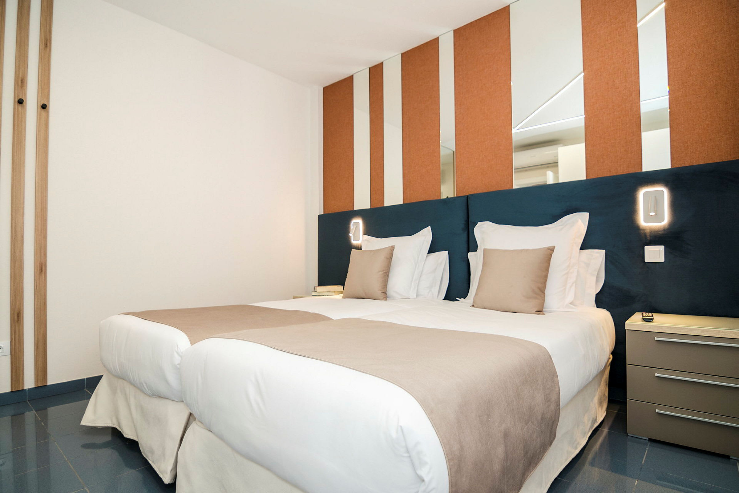 Et moderne hotellrom med to enkeltsenger ved siden av hverandre, oppredt med hvitt og beige sengetøy. Veggene har speil og oransje paneler, og ved siden av sengen er et nattbord. (AI-generert tekst)