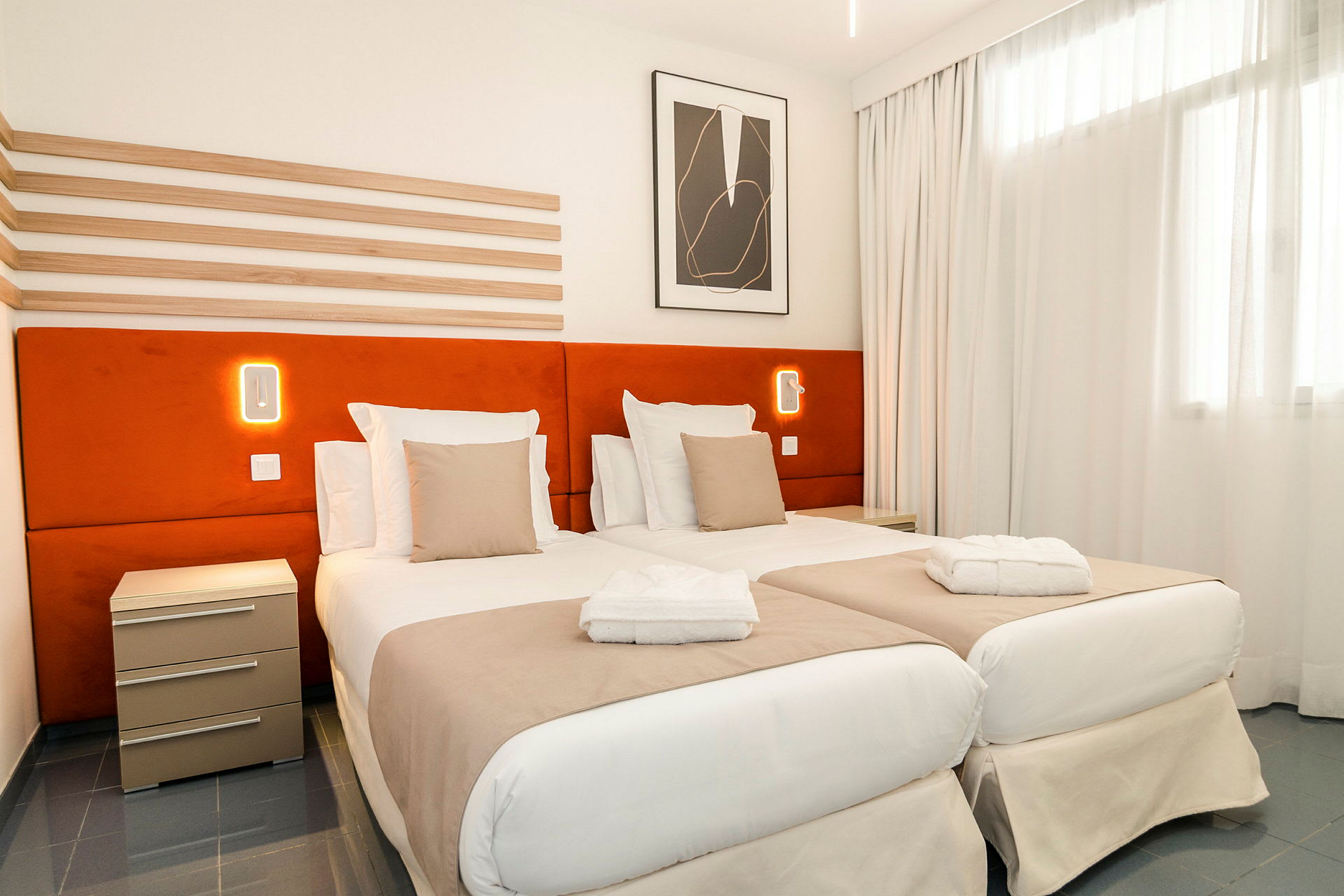 Et hotellrom med to enkeltsenger, oransje sengegavler og hvitt sengetøy. En nattlampe er synlig på veggen. (AI-generert tekst)