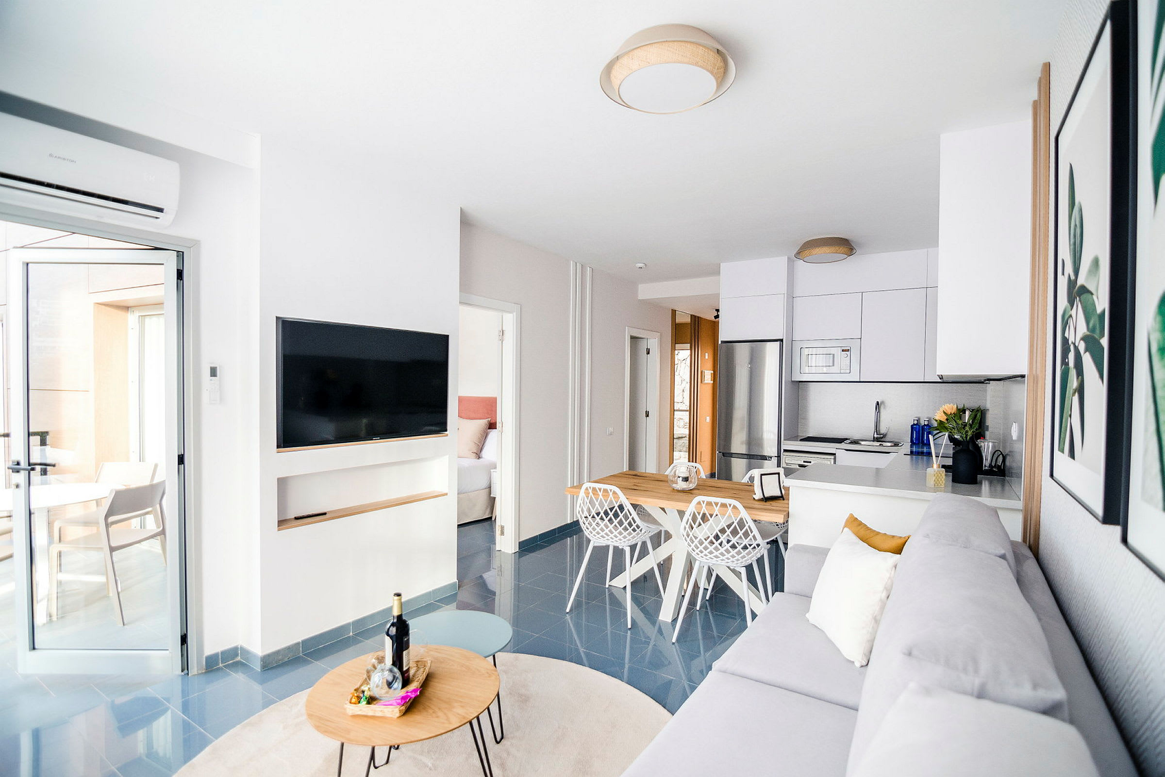En moderne stue med åpen løsning som inkluderer et kjøkken. Rommet har en grå sofa, en flatskjerm-TV på veggen og et spisebord med hvite stoler. En dør fører til et soverom og en annen ut til en lys terrasse. (AI-generert tekst)