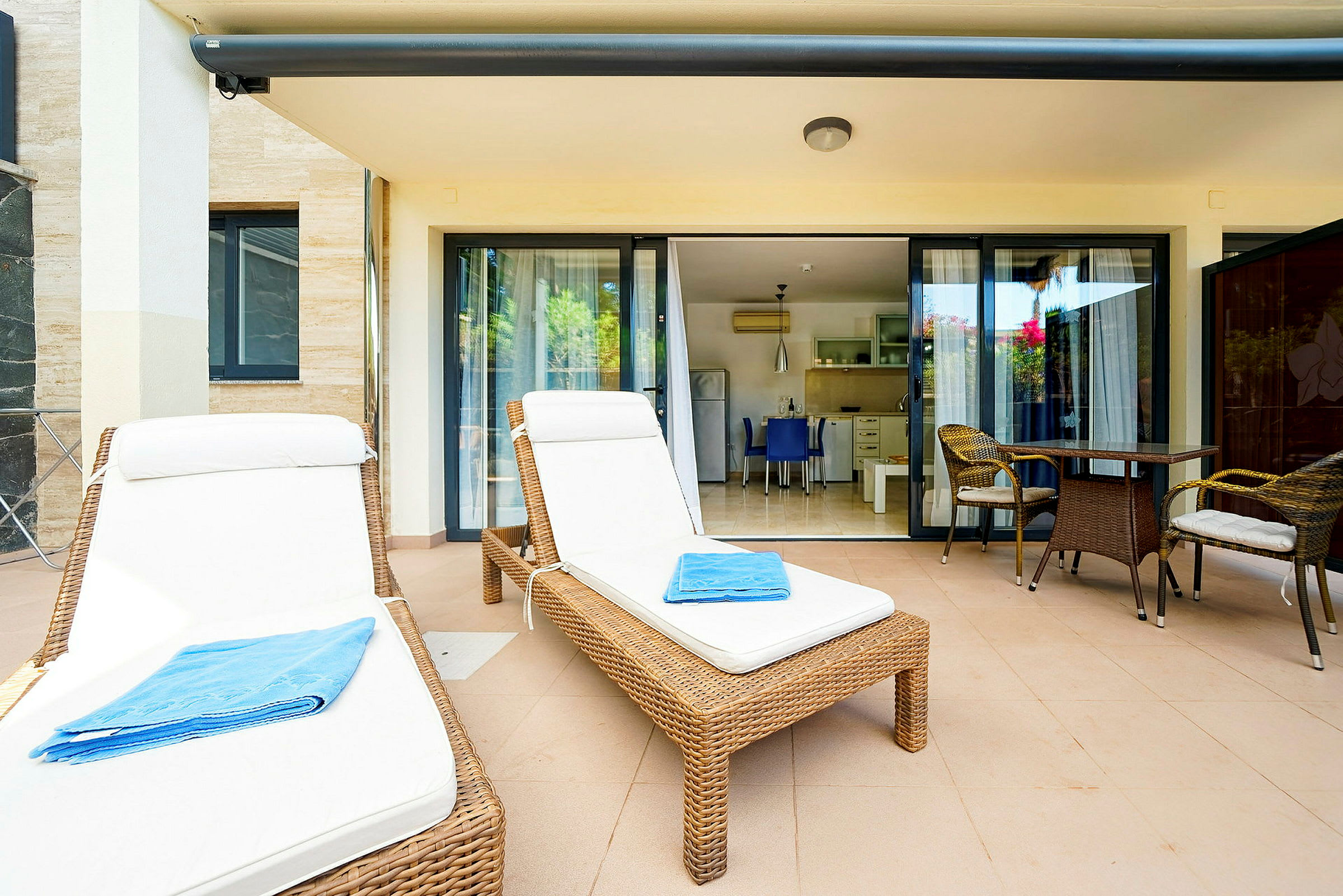 To solsenger med hvite puter og et blått håndkle på en terrasse med bord og stoler foran glassdører til et kjøkken. (AI-generert tekst)