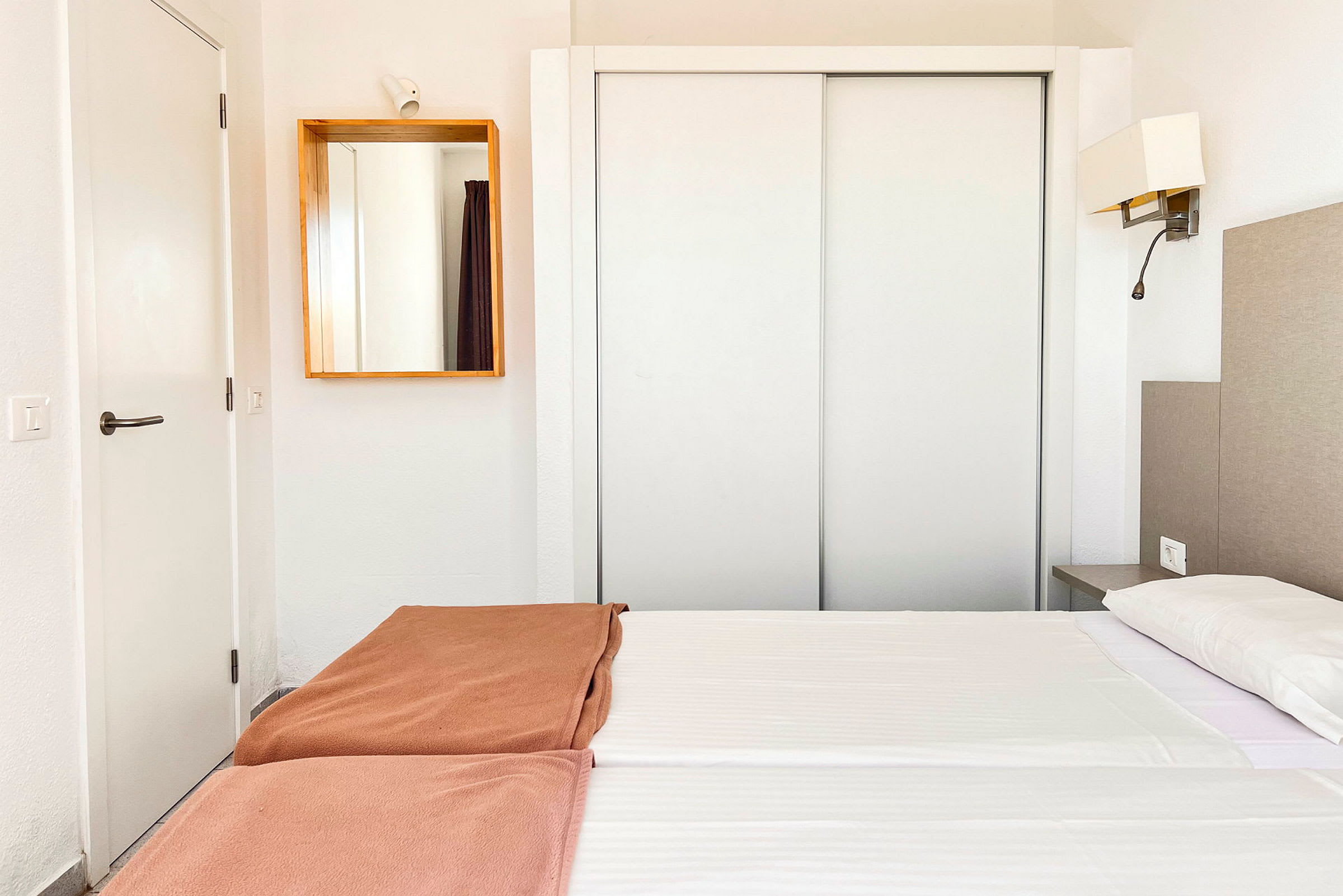 Et hotellrom med hvit innredning, en dobbeltseng med hvite laken og brune sengetepper, samt et speil på veggen. (AI-generert tekst)