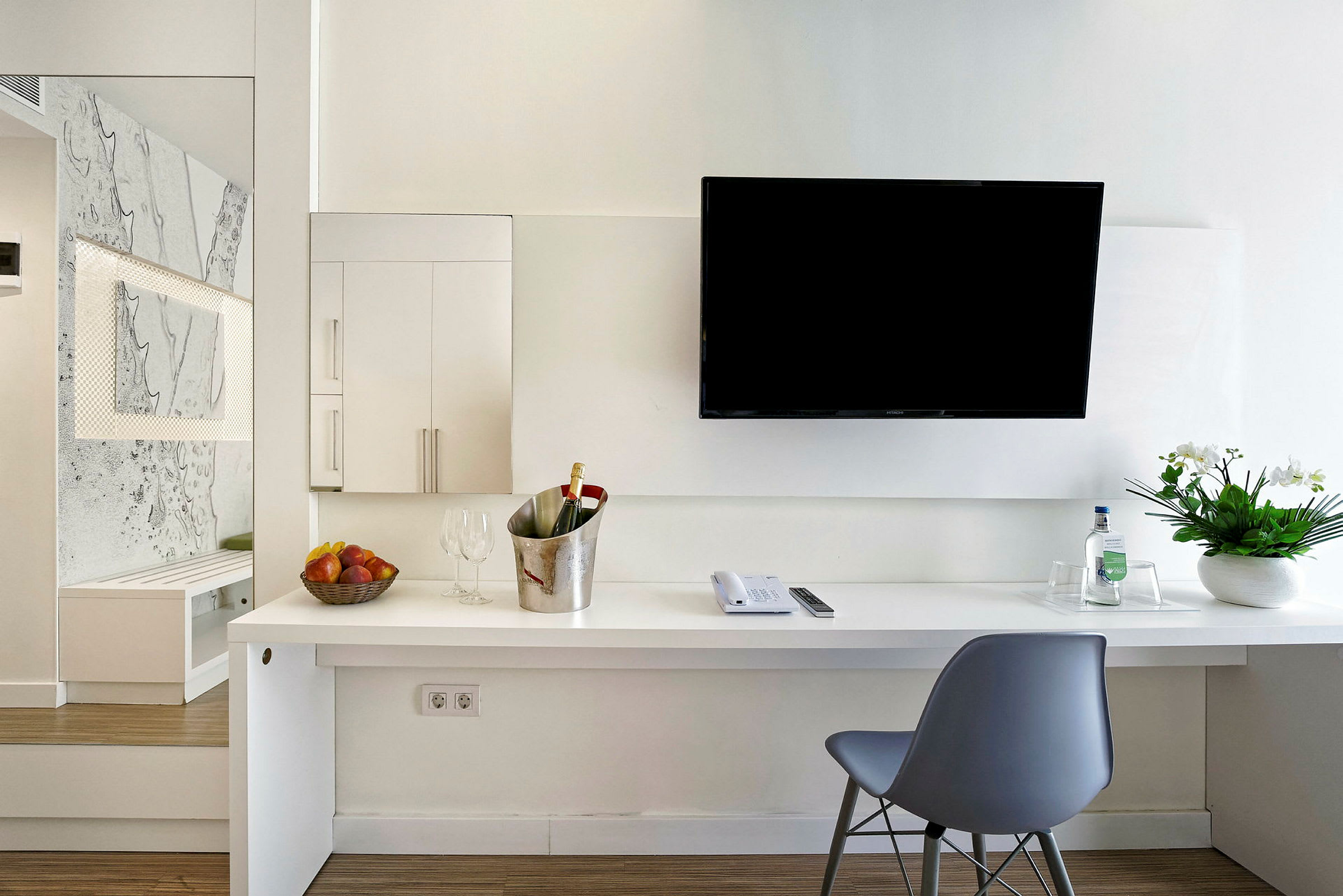 Moderne hotellrominnredning med skrivebord, stol, vegg-tv, blomsterarrangement og forfriskninger. (AI-generert tekst)