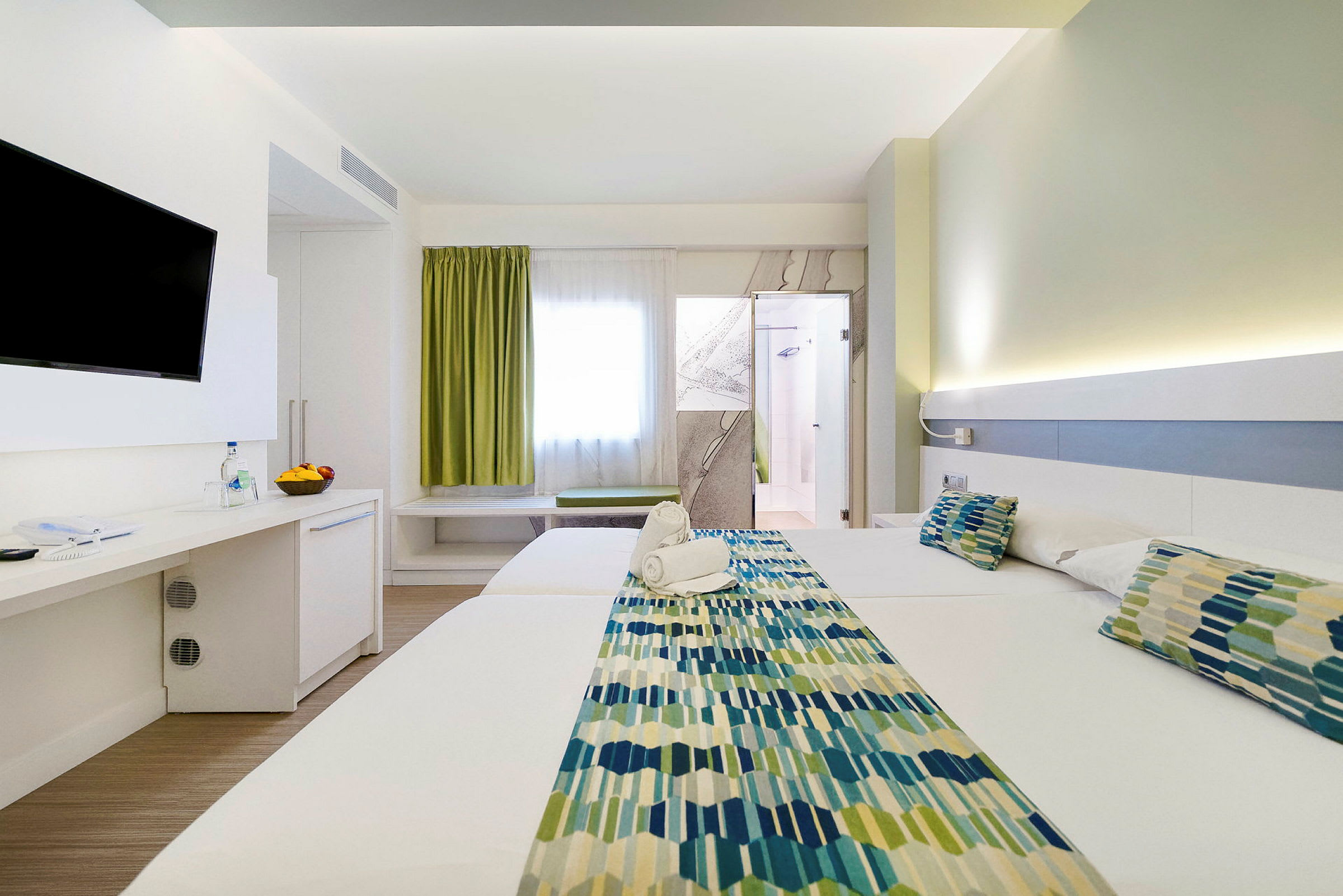 Et hotellrom med to senger, en TV på veggen, og en dør til badet. Rommet har lys innredning. (AI-generert tekst)