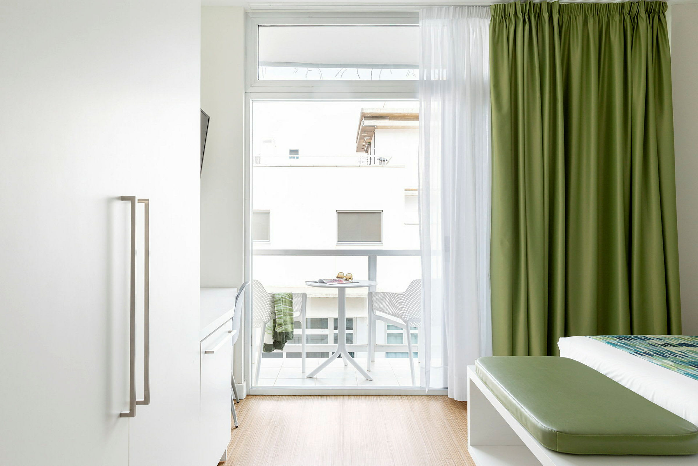 Et lyst hotellrominteriør med en seng, grønne gardiner og en balkong med bord og stoler. (AI-generert tekst)