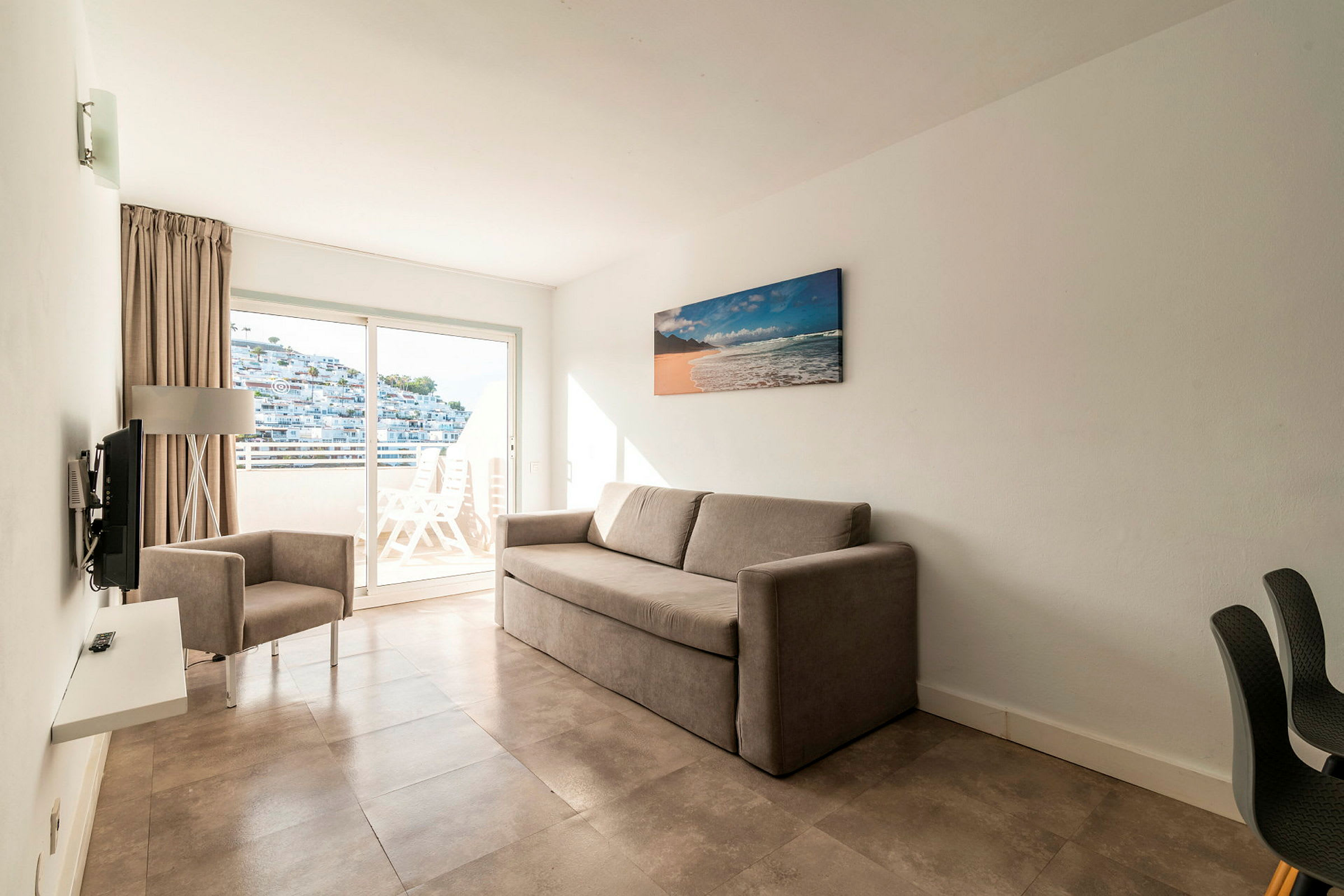 Et hotellrom med en beige sofa og en lenestol samt en balkong med utsikt. (AI-generert tekst)