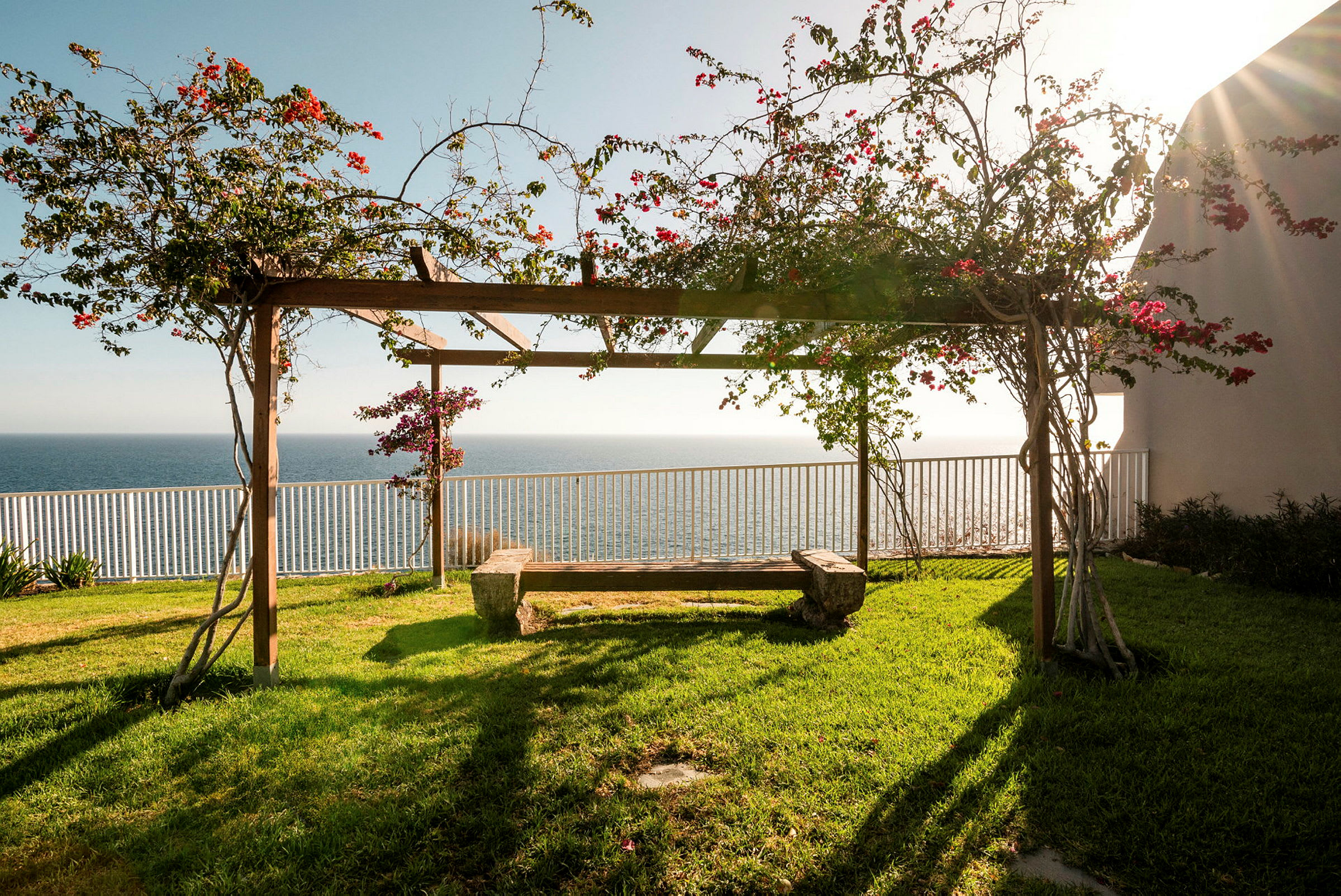 En pergola med blomstrende planter og benk, havet i bakgrunnen. (AI-generert tekst)