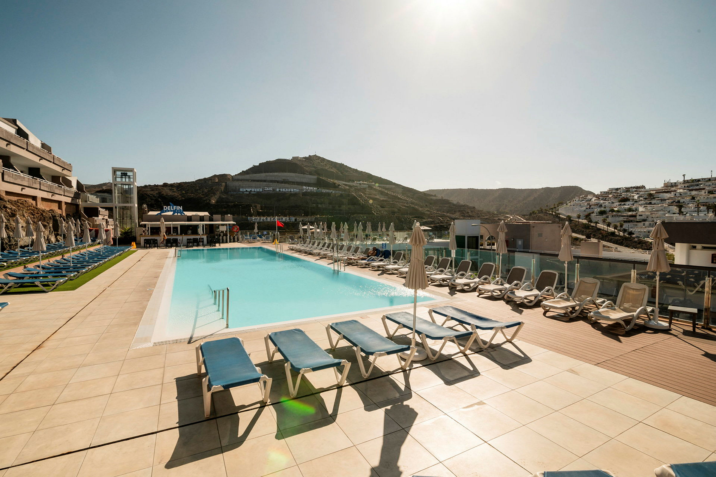 Utendørs utsikt over et hotellbasseng med solsenger og parasoller på et solrikt fjellområde. (AI-generert tekst)