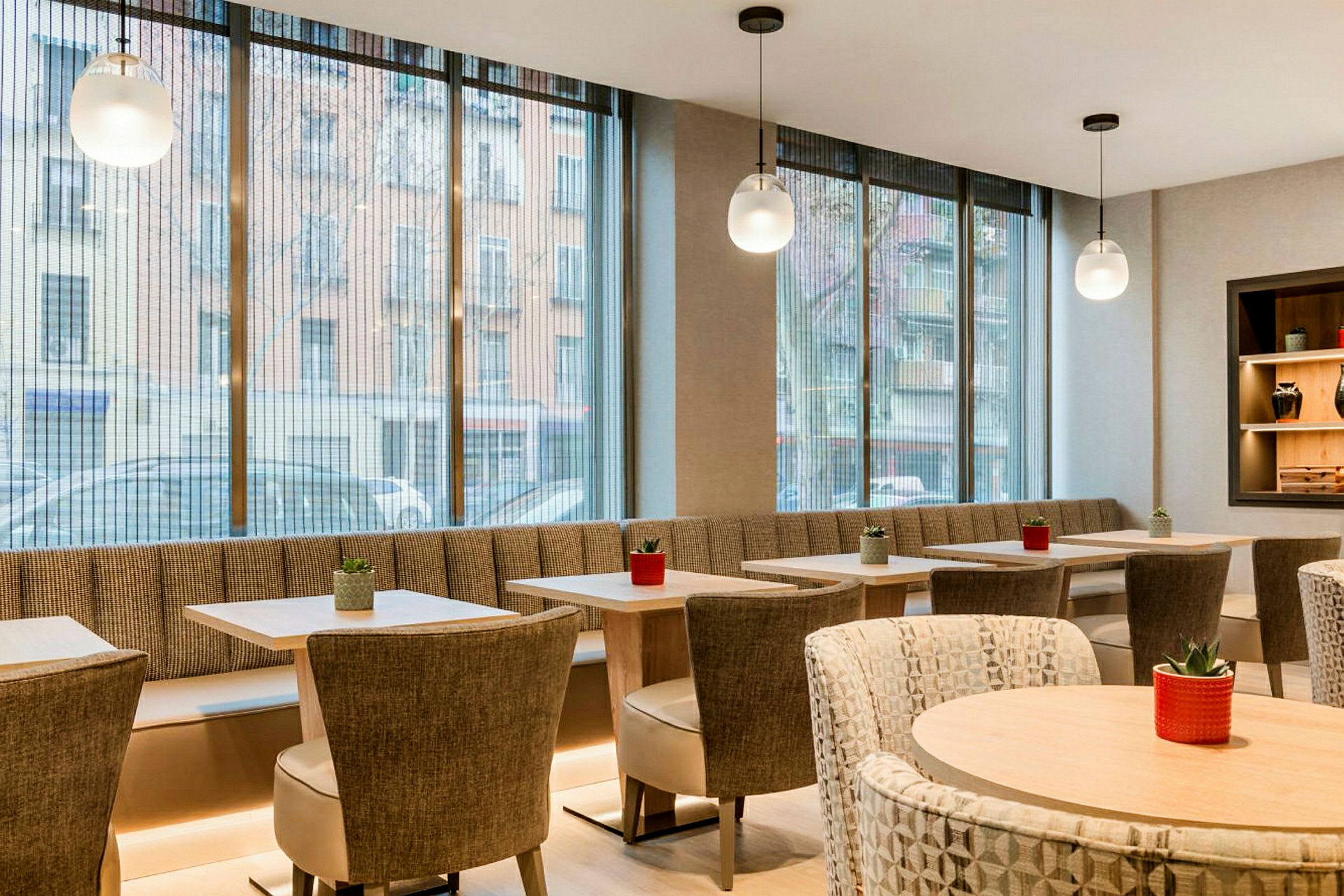 Et moderne og lyst innredet kafé med store glassvinduer, bord og stoler samt små planter som dekorasjon. (AI-generert tekst)