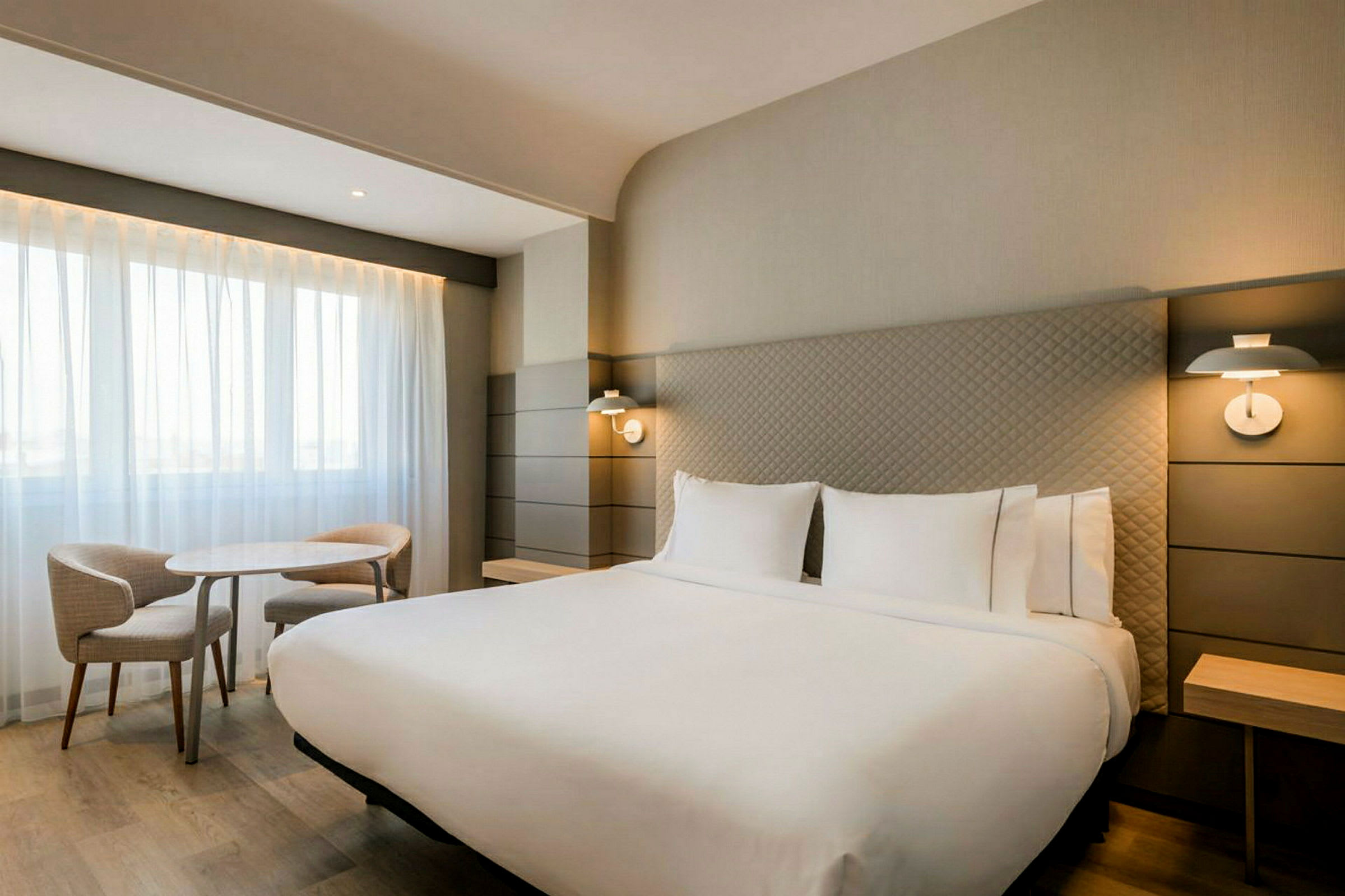 Et moderne hotellrom med en stor dobbeltseng, to sengelamper, et bord og to stoler ved et vindu med lyse gardiner. (AI-generert tekst)