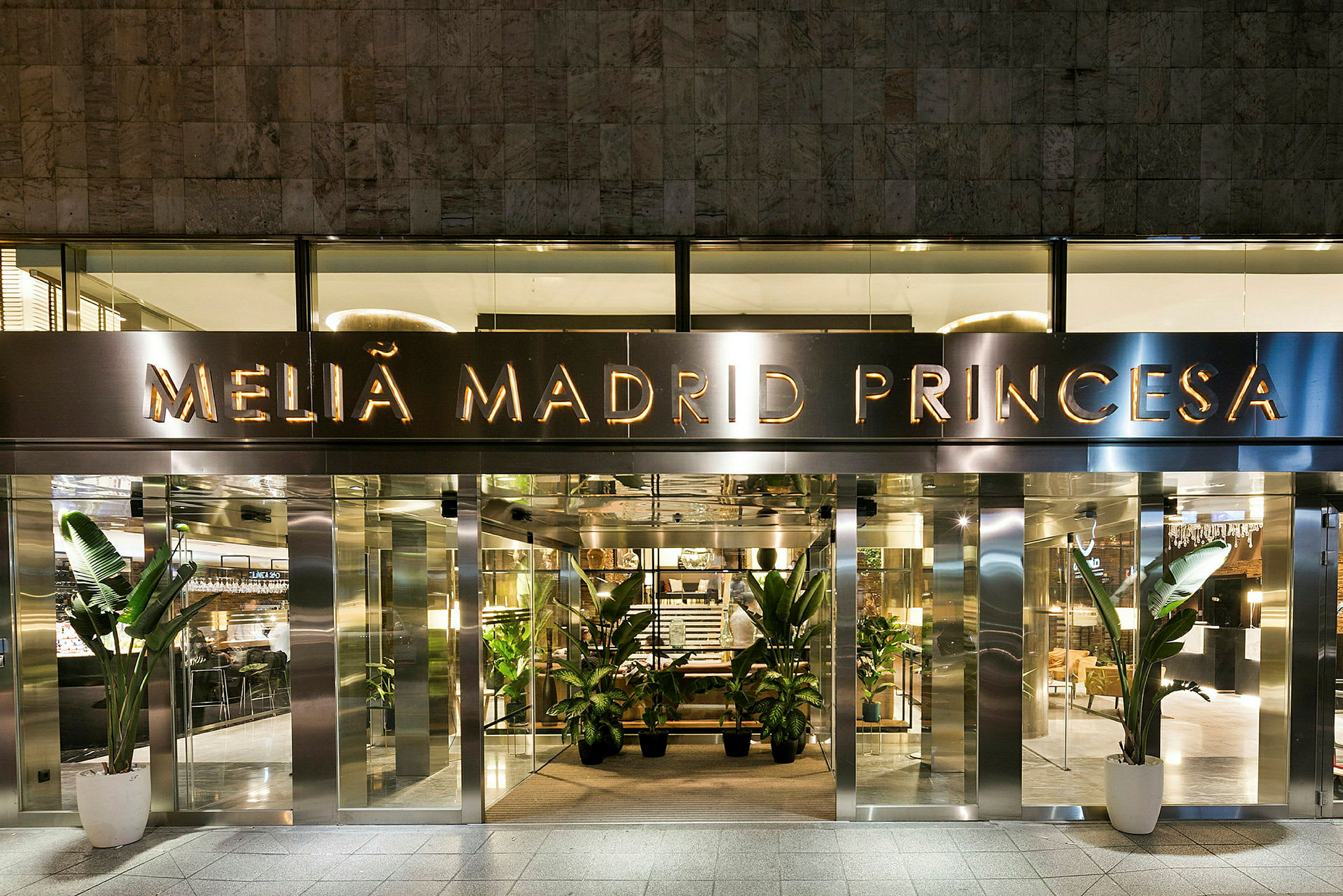 Inngangen til hotellet Melia Madrid Princesa, med glassdører og dekorative planter ved inngangen. (AI-generert tekst)