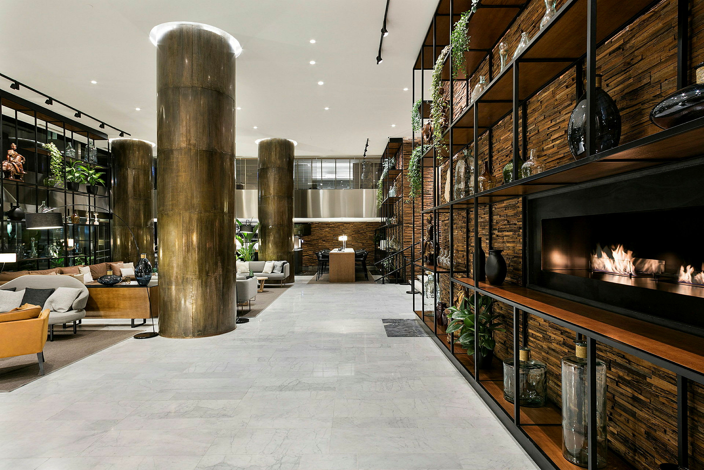 Et moderne hotellobbyområde med sitteplasser, marmorgulv, store søyler og en peis innebygd i en hyllevegg. (AI-generert tekst)