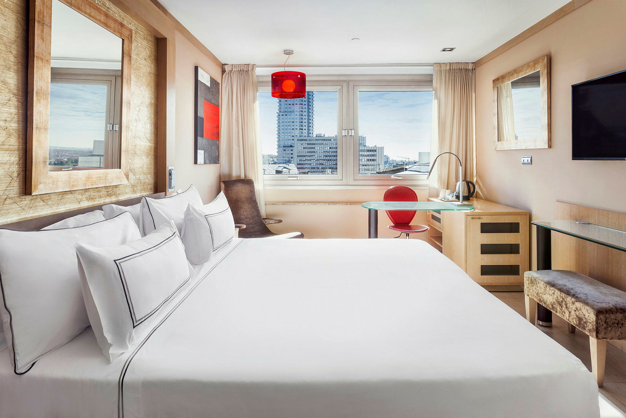Et lyst og moderne hotellrom med stor seng, vindu med utsikt over byens bygninger, et skrivebord med rød stol og en veggmontert TV. (AI-generert tekst)