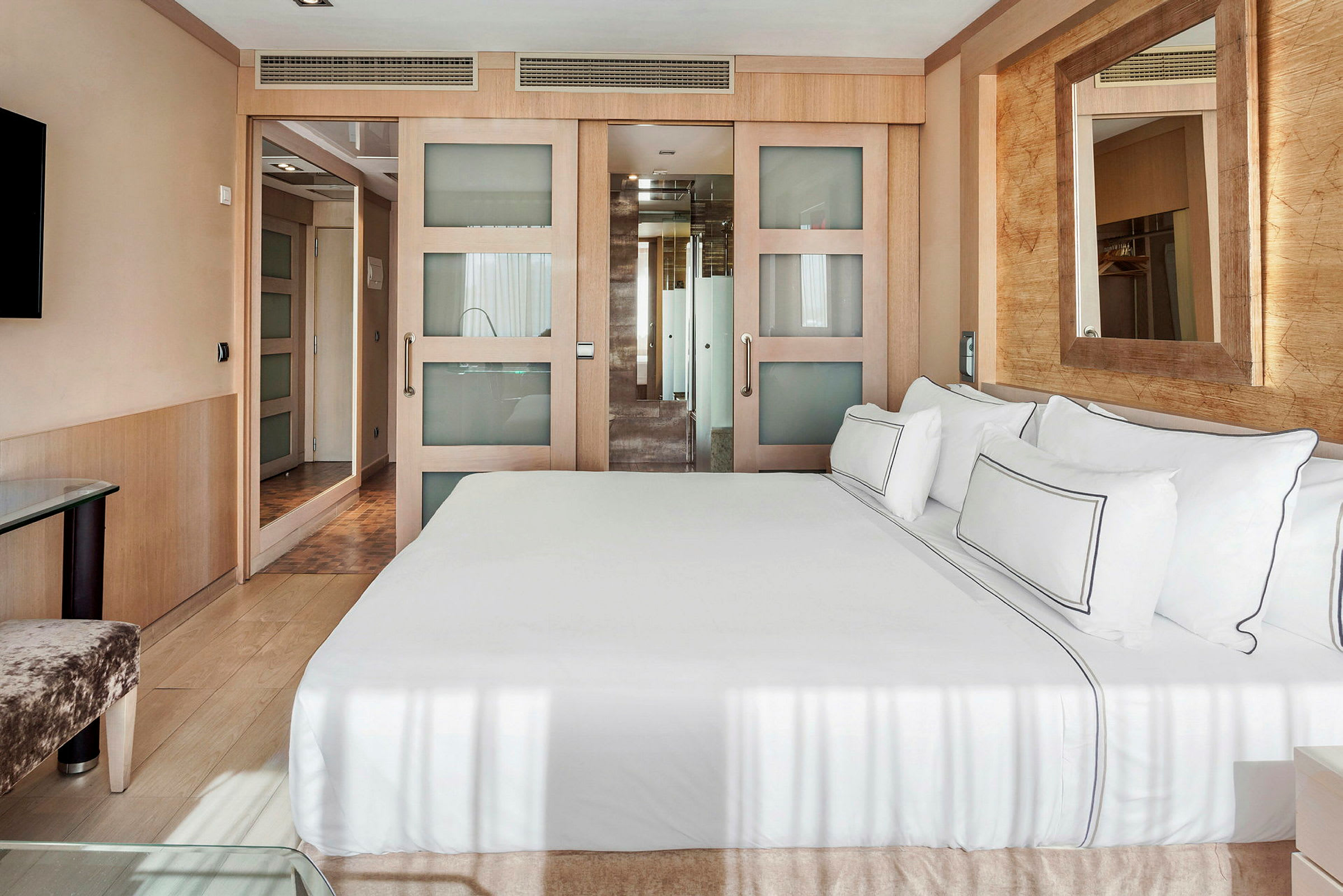Et moderne hotellrom med en stor seng utstyrt med hvitt sengetøy, et speil på veggen, og en skyvedør som leder til et bad. (AI-generert tekst)